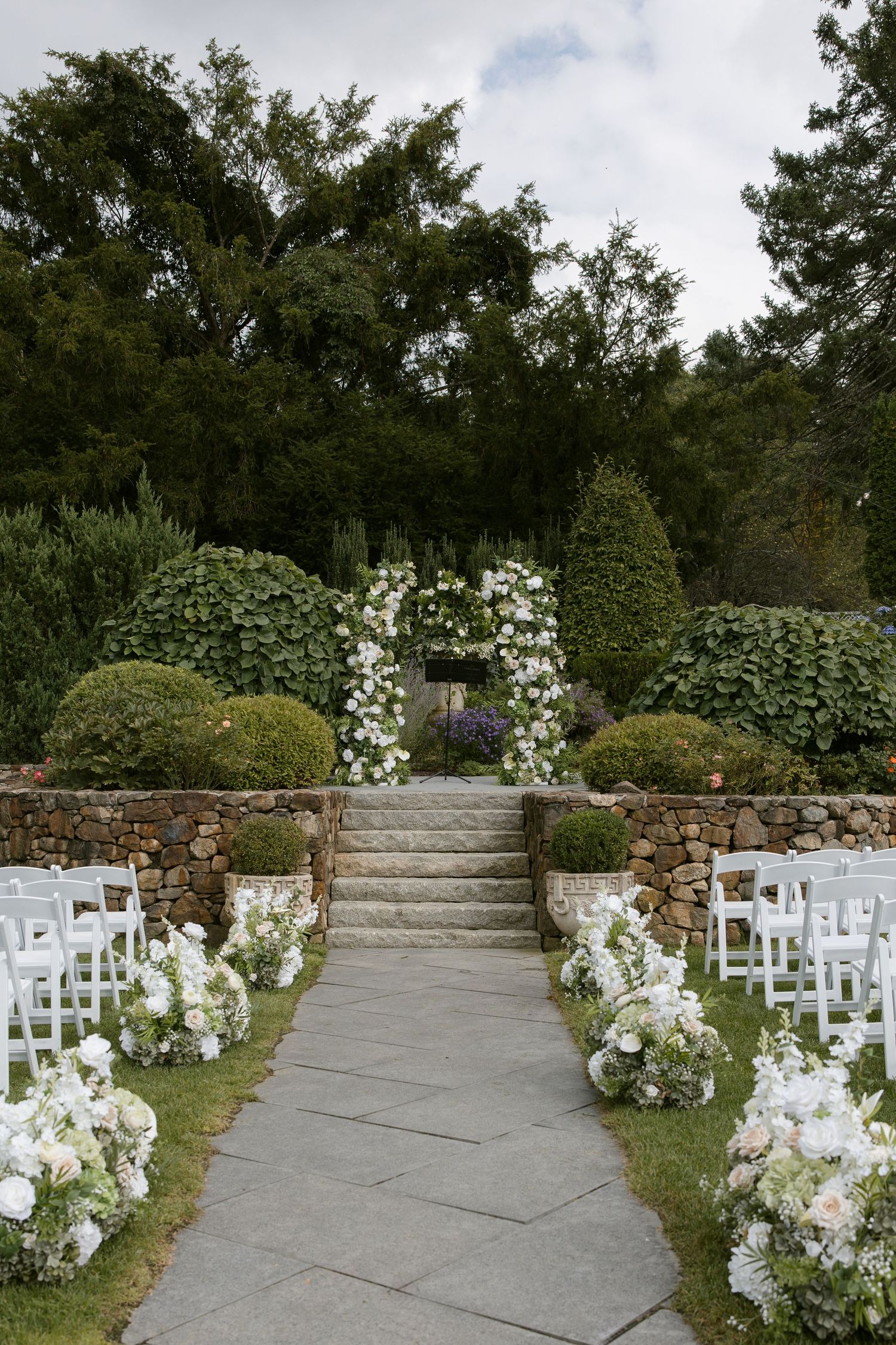 Elegant outdoor wedding ceremony aisle with white floral arrangements leading to a floral arch and stone steps.