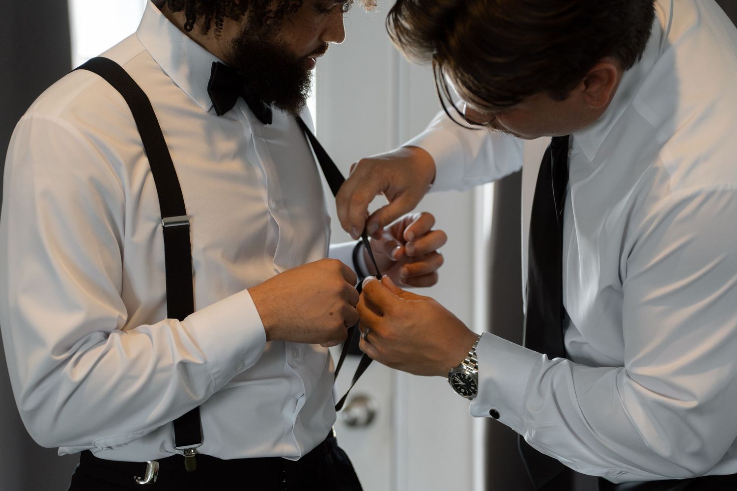 Two groomsmen in white shirts and suspenders helping each other with a tie before a wedding ceremony.