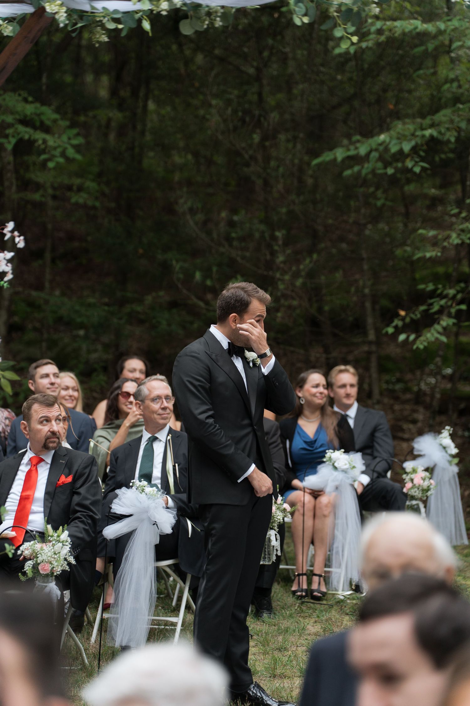 Emotional groom covers face during outdoor wedding ceremony while guests smile in background.