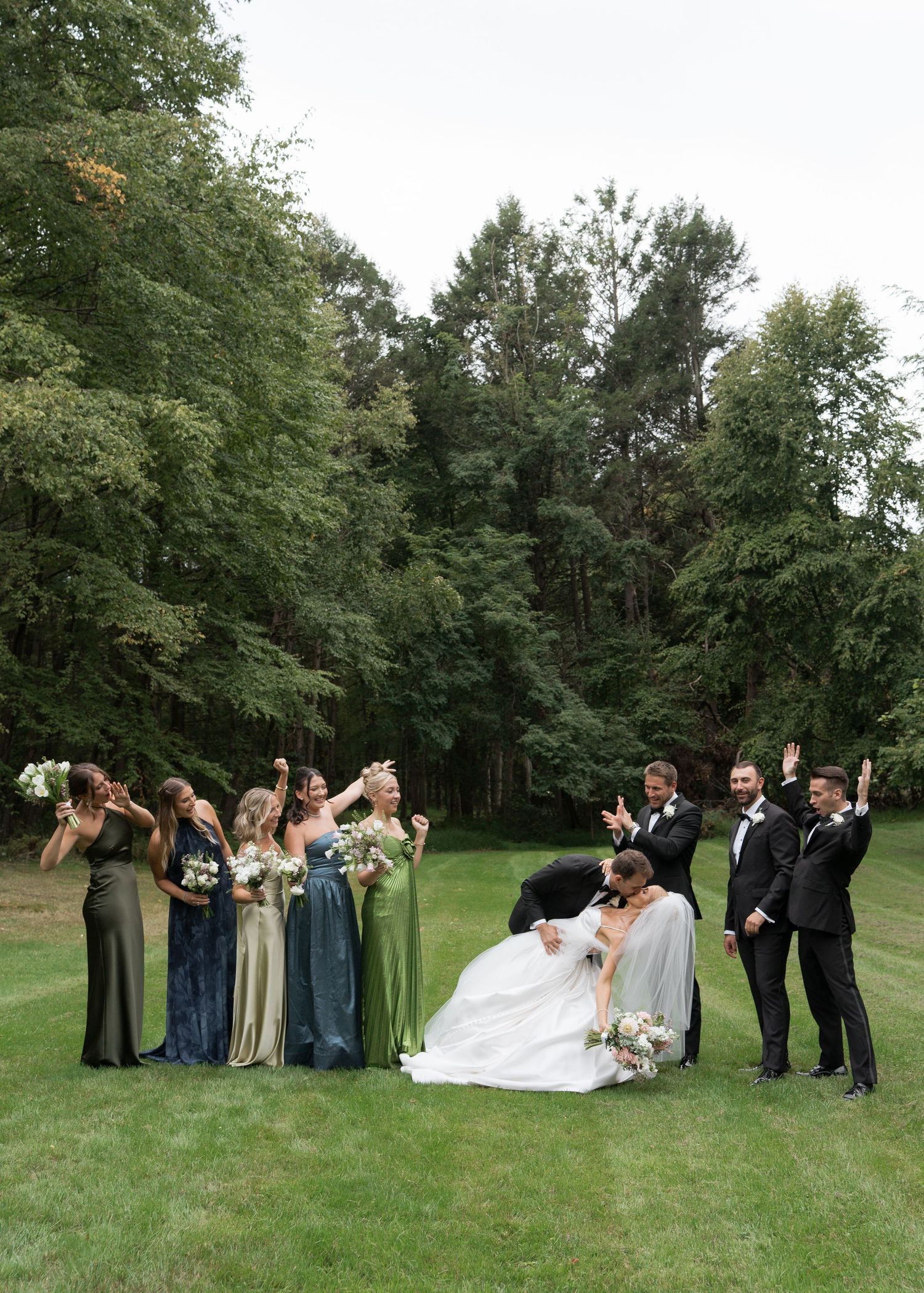 Wedding party celebrating on green lawn with bridesmaids in sage dresses and groomsmen in tuxedos.