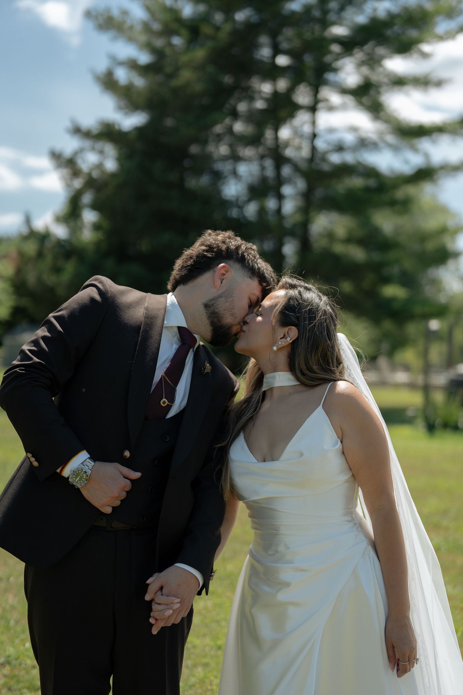 Bride and groom sharing a kiss outdoors on their wedding day, with lush green trees in the background.