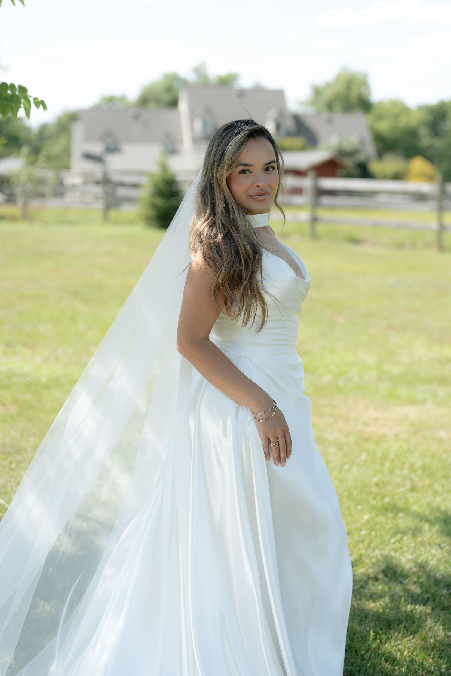 Bride in white satin gown and flowing veil poses outdoors on green grass with rustic fence and farmhouse backdrop.