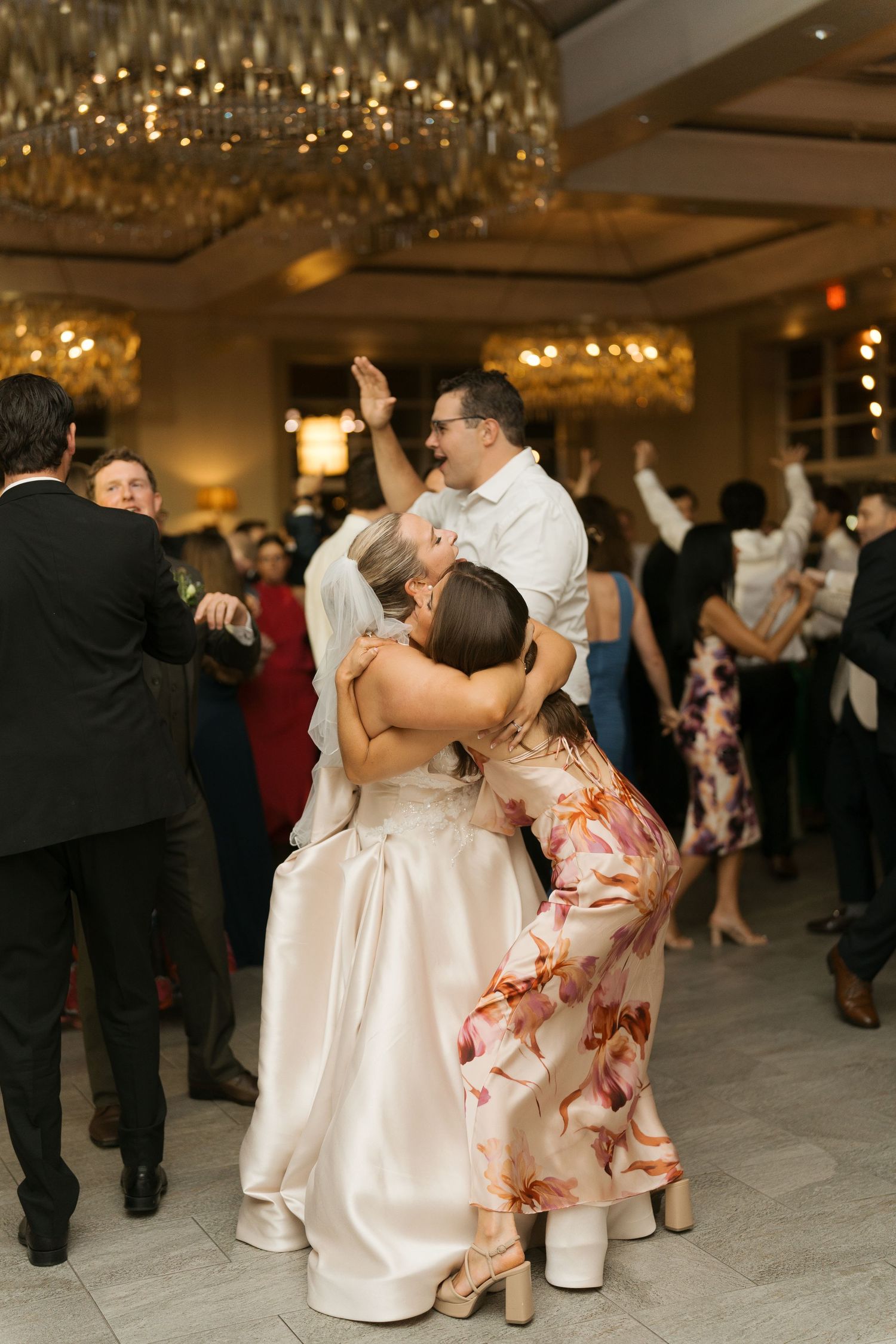Bride in white satin gown joyfully embraced by guests on a crowded wedding reception dance floor under a chandelier.