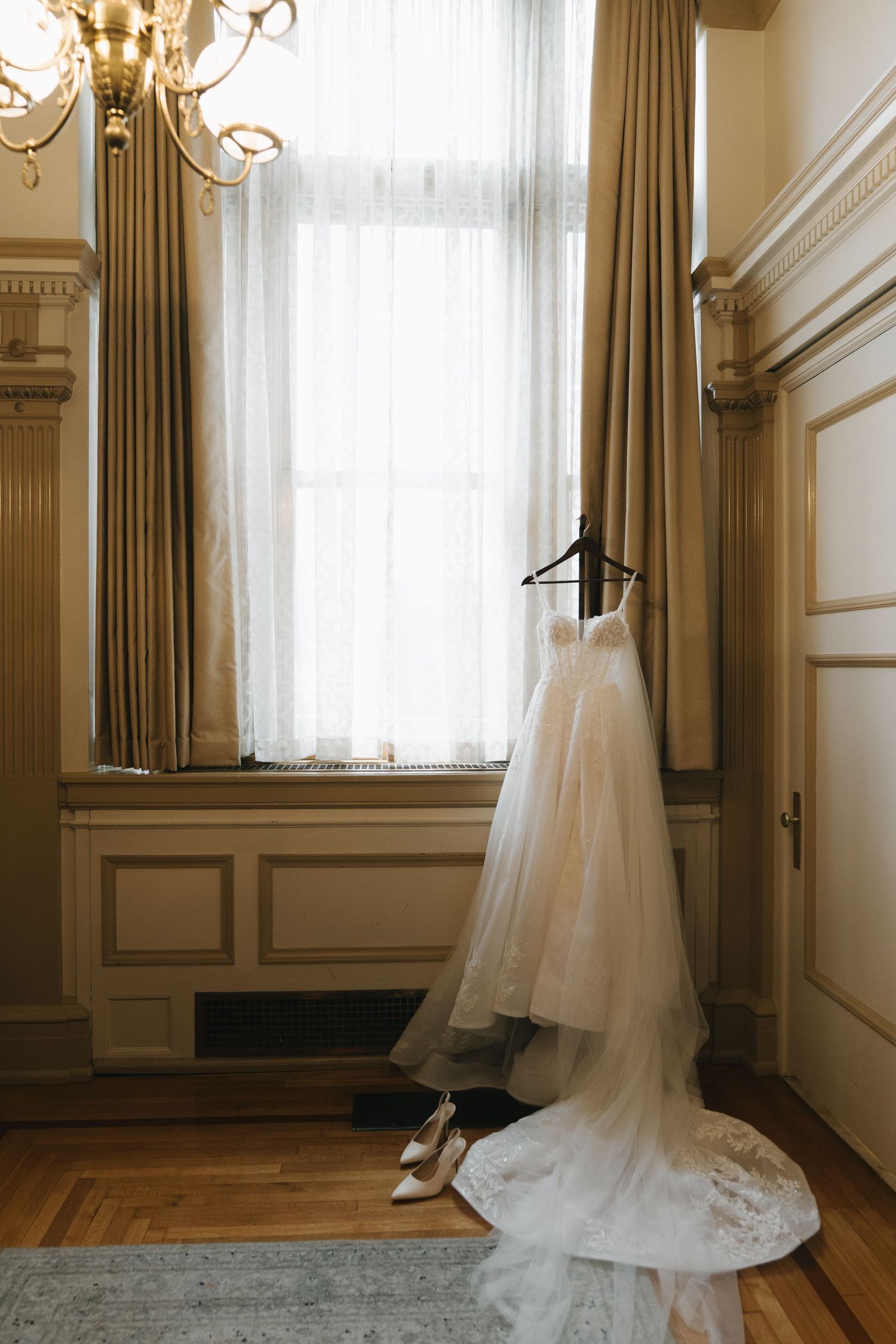 Elegant wedding dress hanging by a sunlit window with bridal heels below in a classic wood-paneled room.