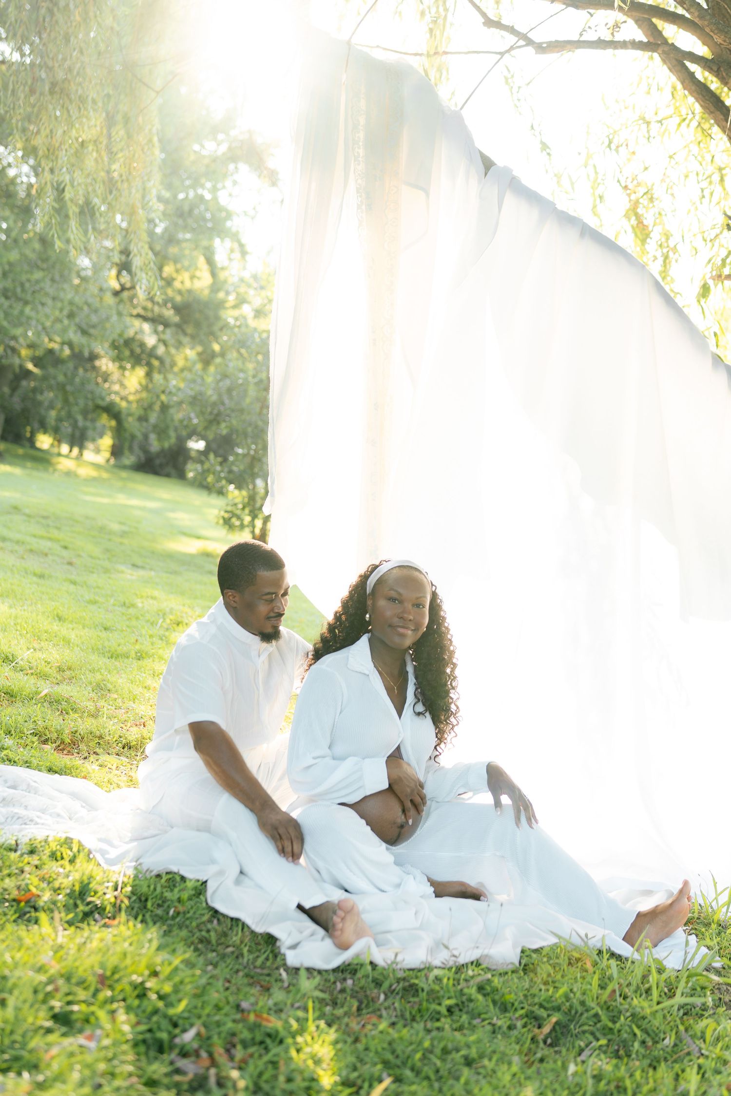 A pregnant couple in white outfits sits on grass in sunlit park, with sheer white fabric backdrop behind them.