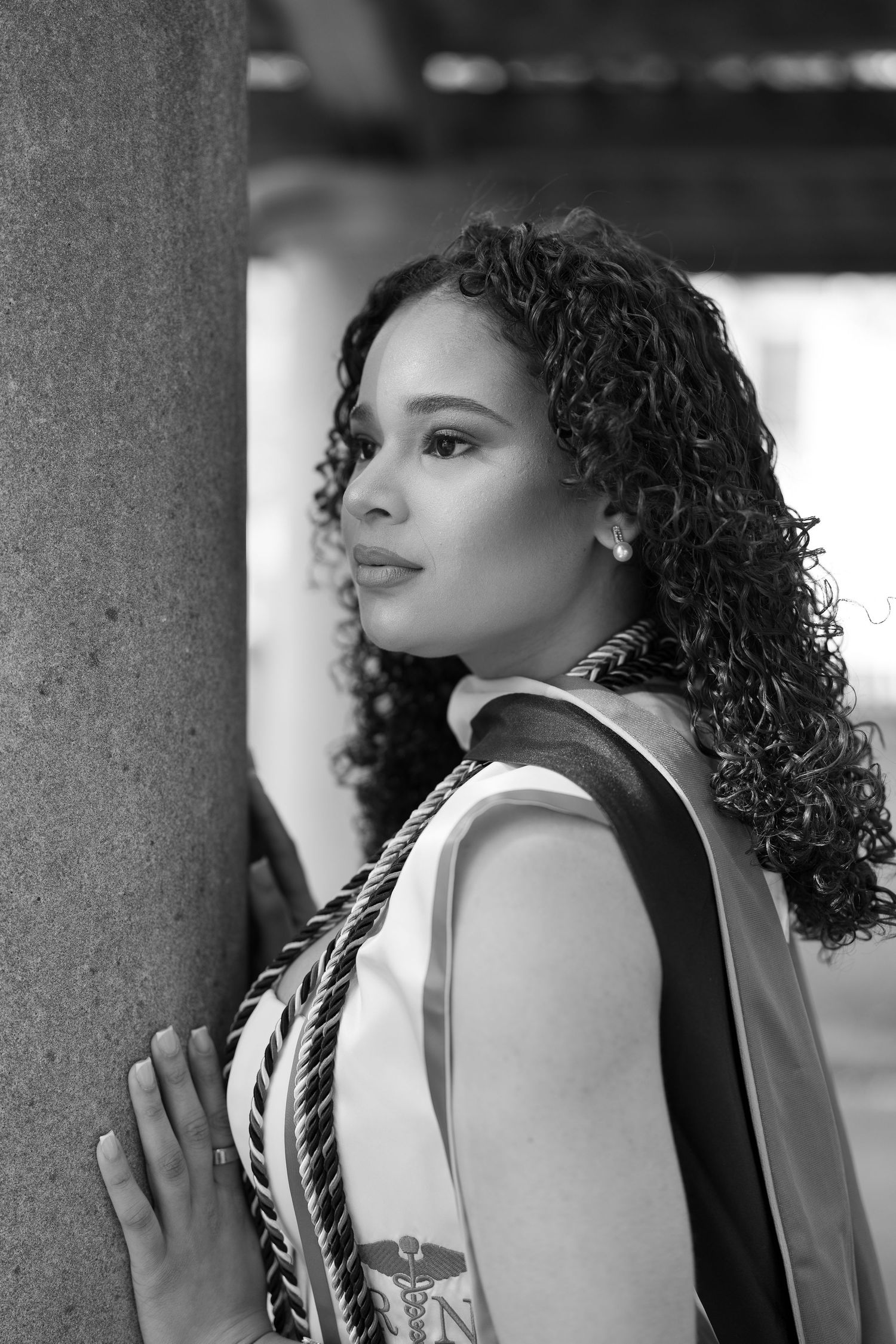 Black and white portrait of curly-haired woman leaning against stone column wearing graduation sash.