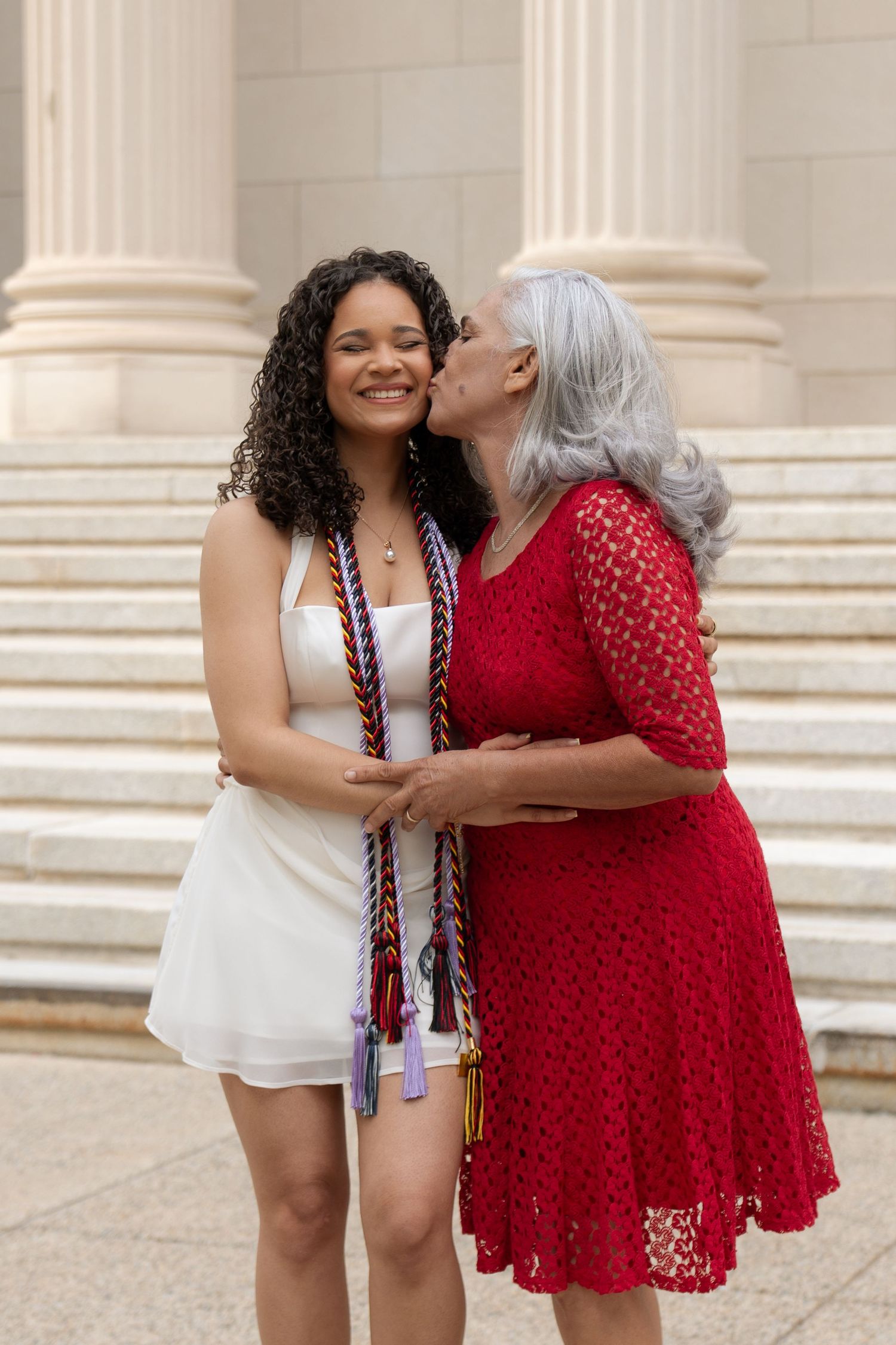 Older woman with gray hair kissing young female nursing graduate wearing colorful honor cords outdoors.