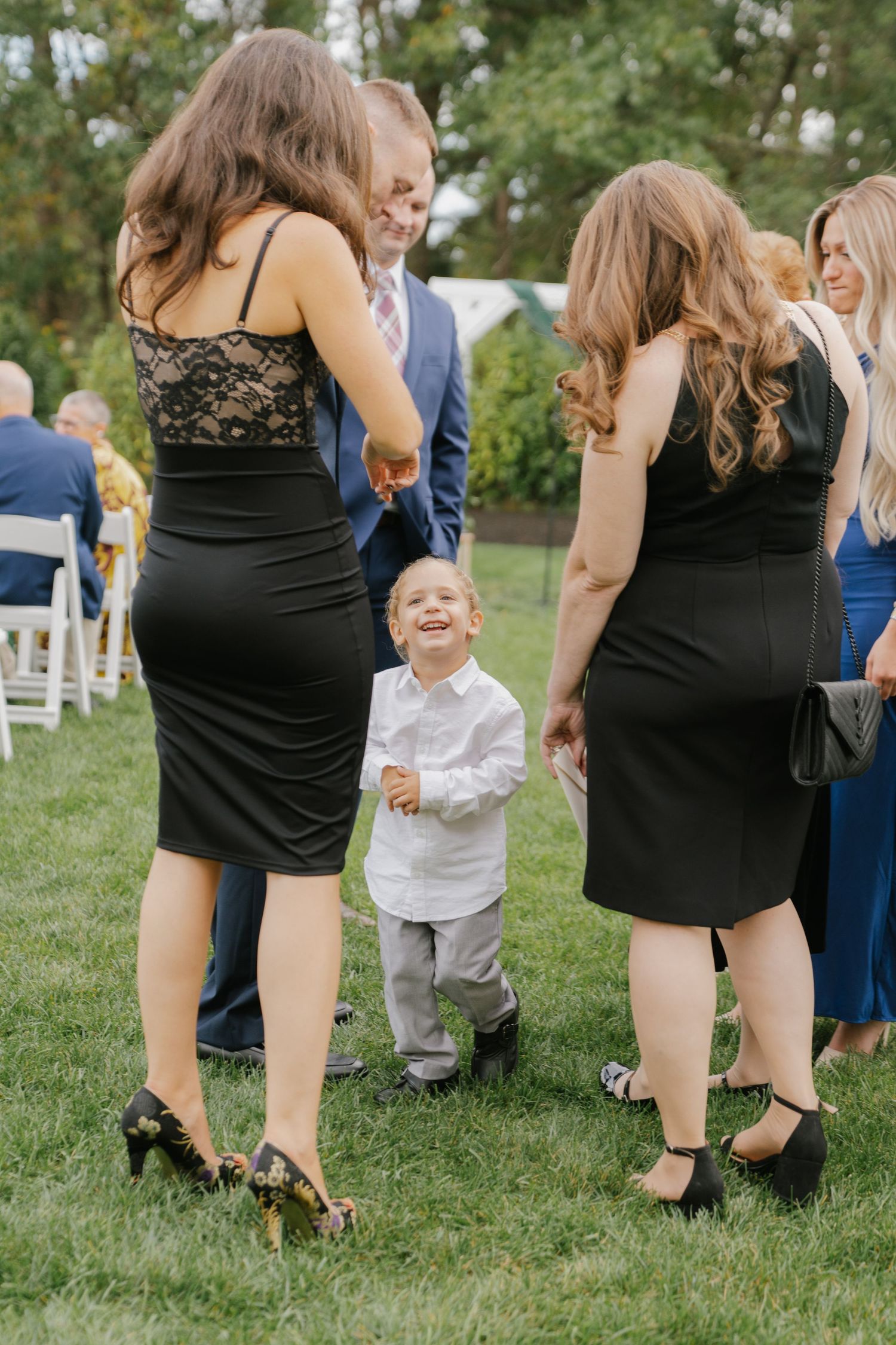 A young child in a white shirt laughs joyfully between two women in black dresses at an outdoor wedding ceremony.