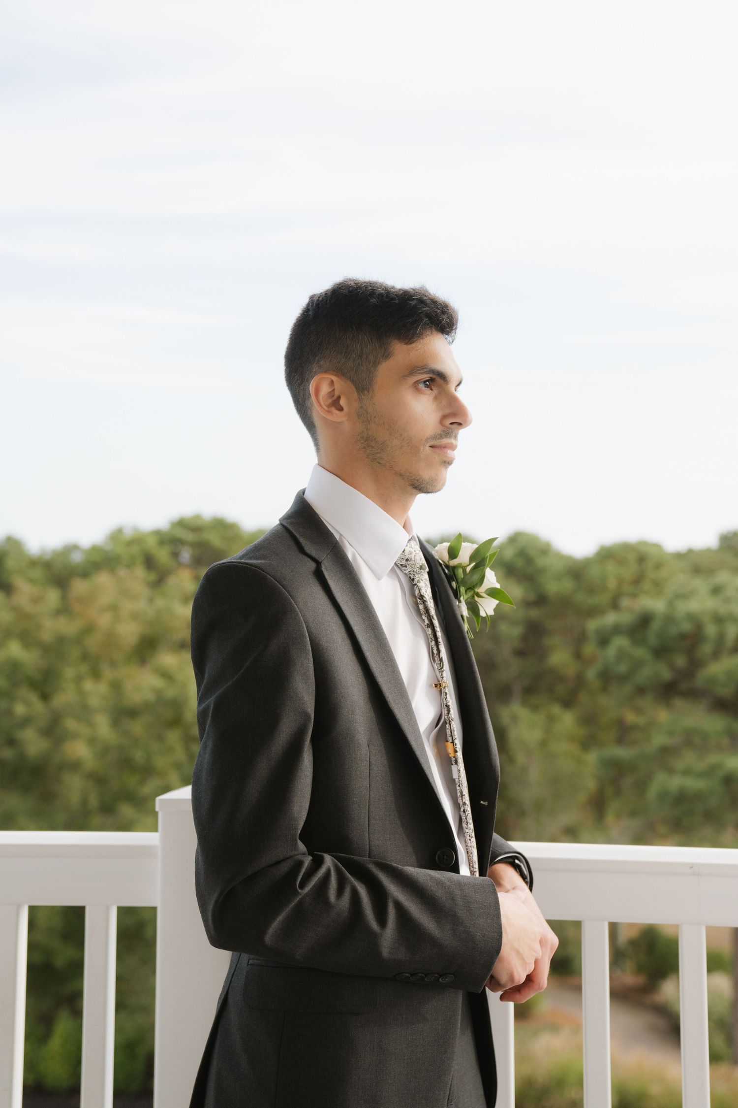 A groom in a black suit stands thoughtfully on a white railing deck overlooking a scenic natural landscape.