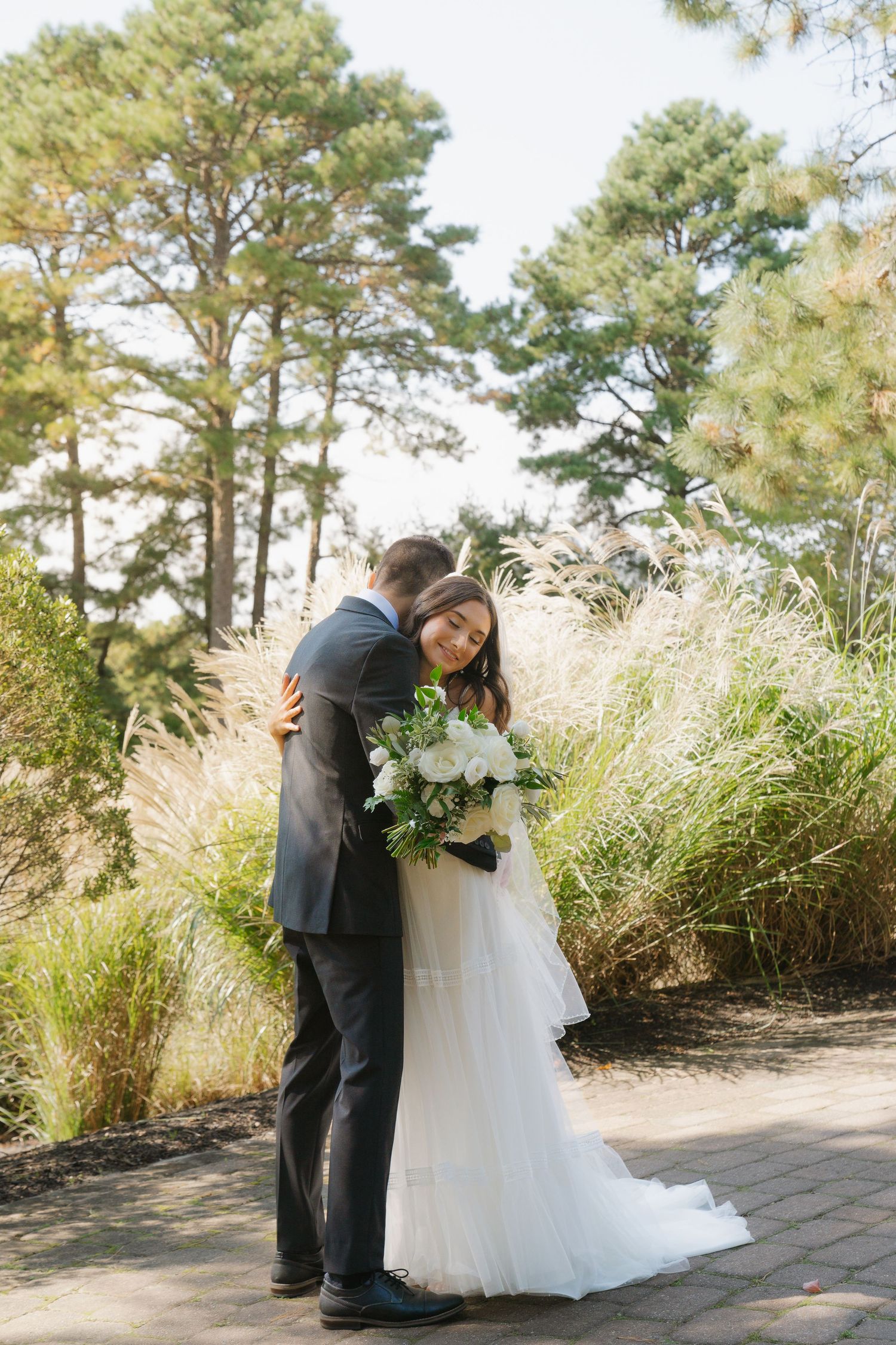 A couple shares an intimate moment surrounded by tall pine trees and ornamental grasses on a stone pathway.
