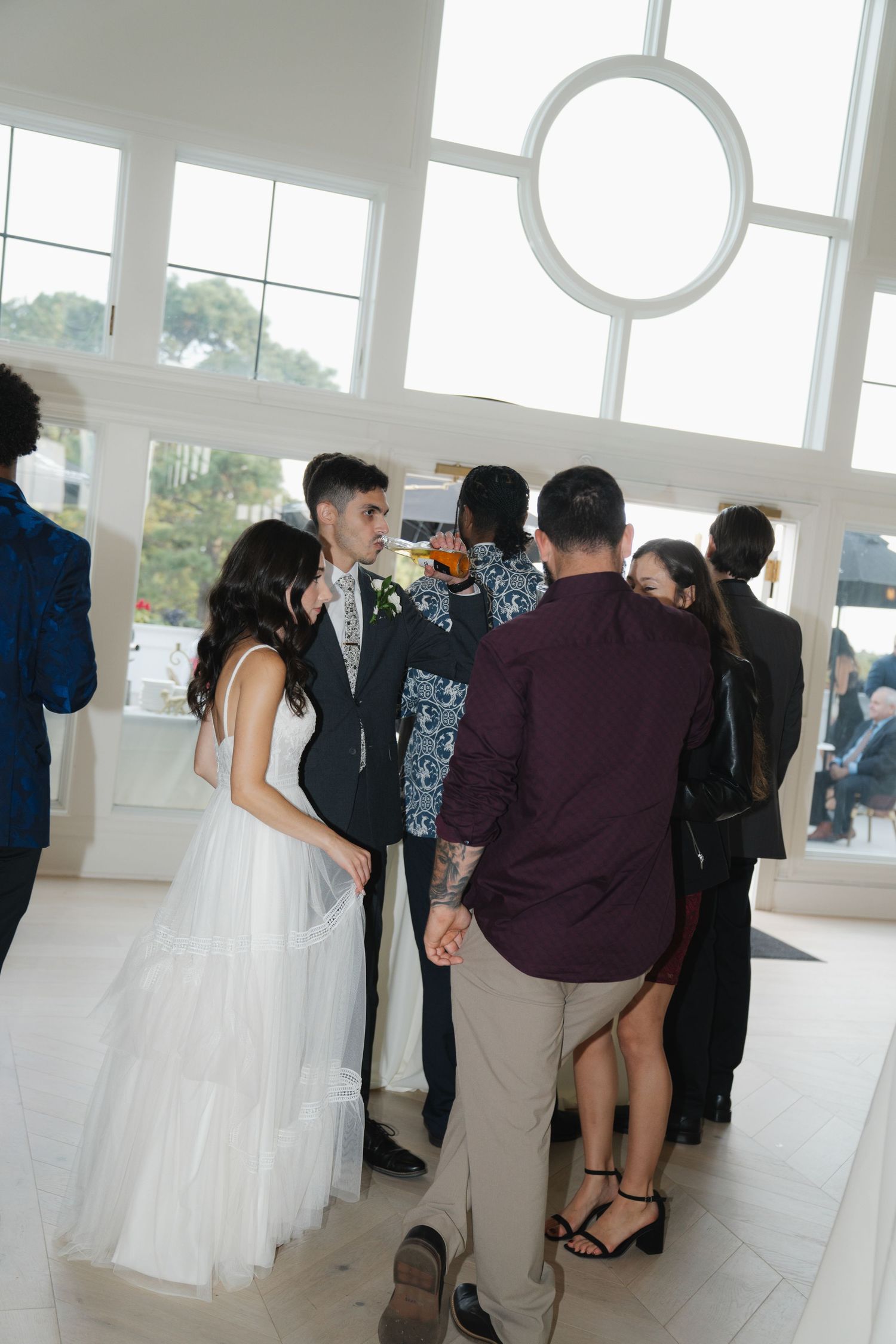 Wedding guests mingle and socialize in a bright, modern venue with large windows and white decor.