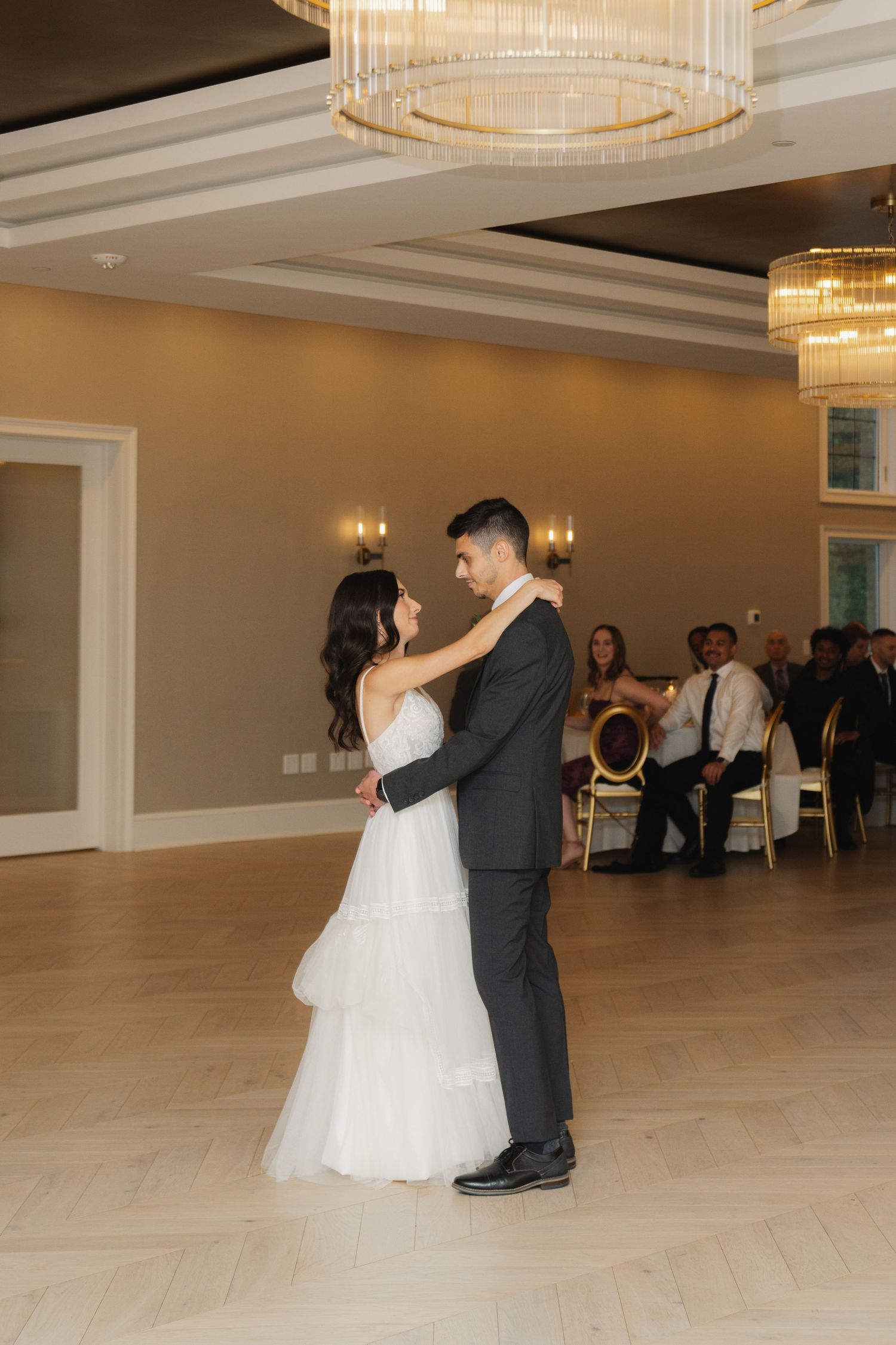 A bride and groom share a romantic dance on a light wood floor under crystal chandeliers at their wedding reception.