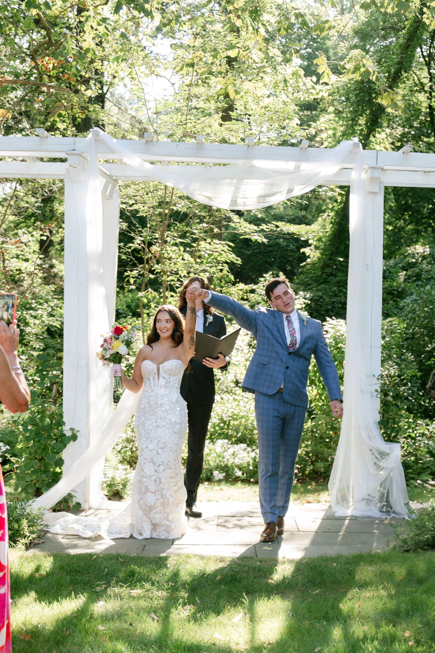 Celebration sequence under a white pergola with greenery and sunlight filtering through.