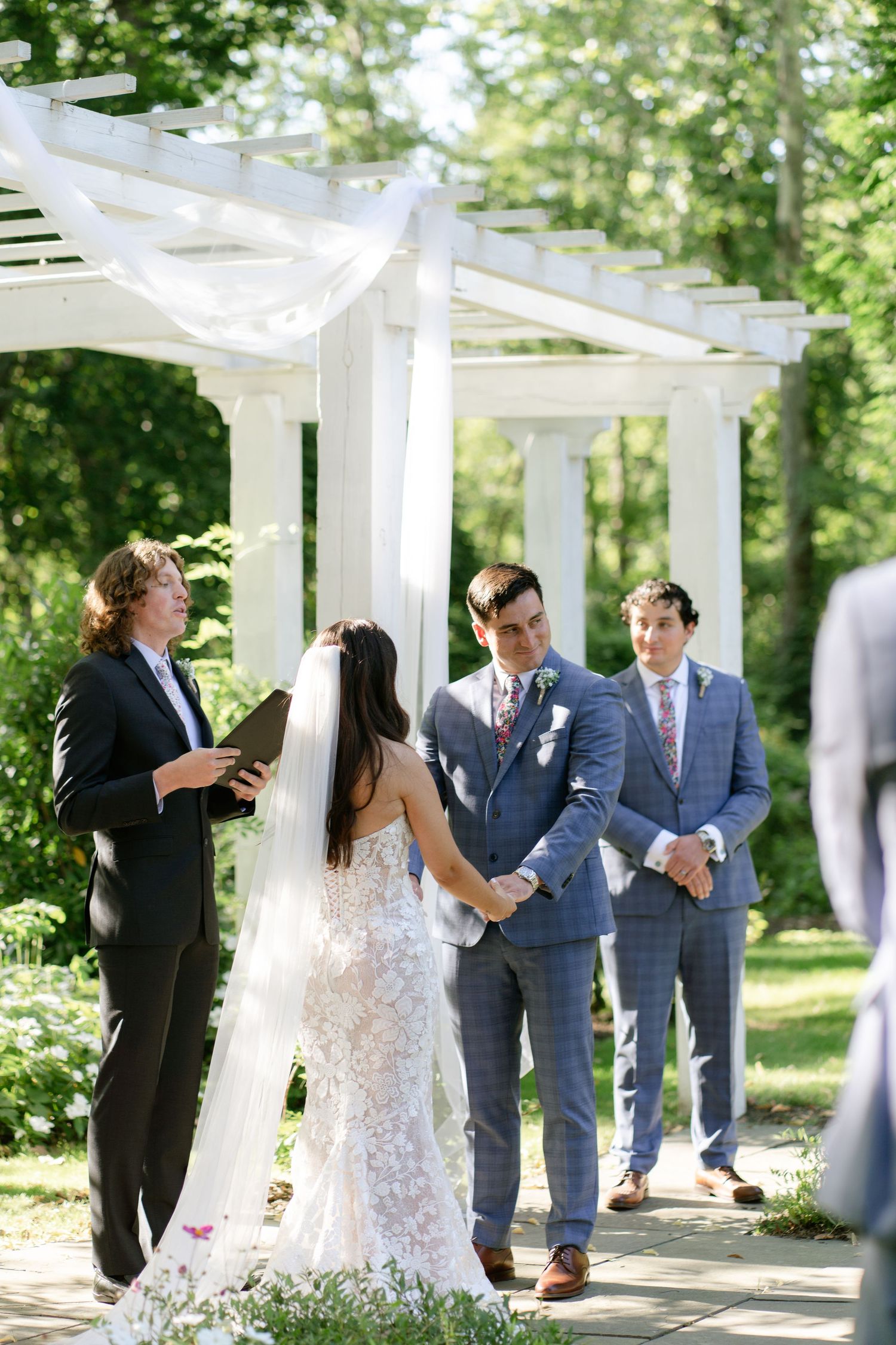 Wedding ceremony taking place under a white pergola in a sunny garden setting.