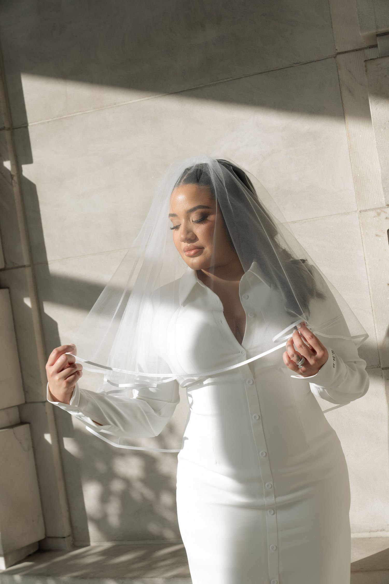 A bride in a sleek white wedding dress poses elegantly near a sunlit window.