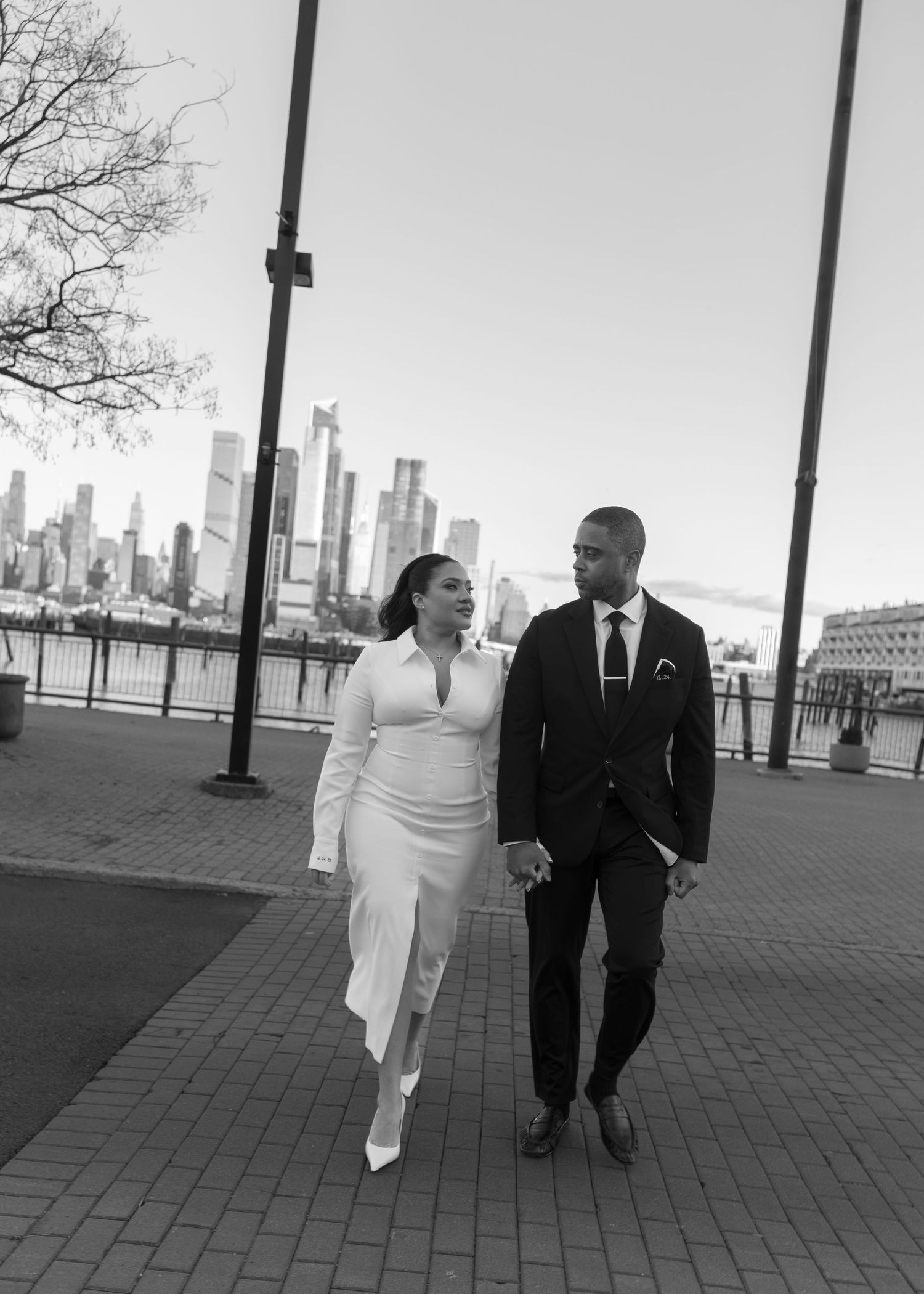 A newlywed couple walks together along a waterfront promenade with city skyline in the background.