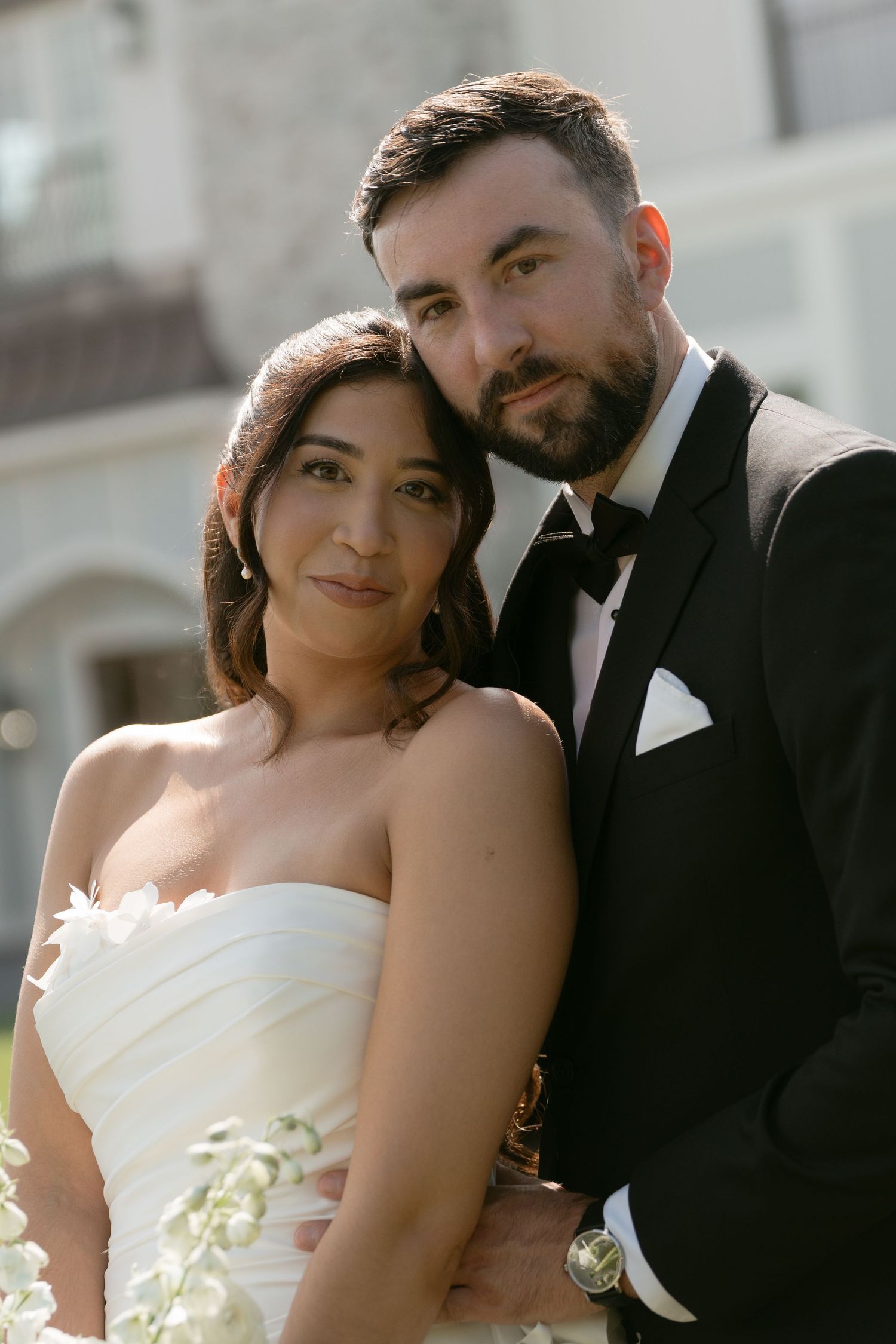 Close portrait of bride in strapless white dress and bearded groom in black tuxedo posing together outdoors.
