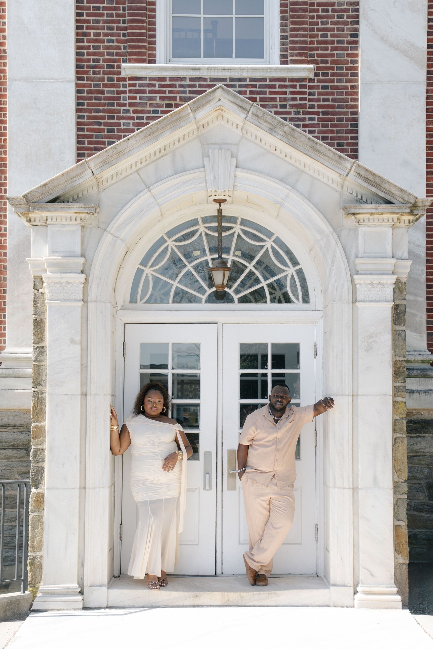 Couple in white and beige outfits smiling at elegant brick building entrance with arched white doorway.