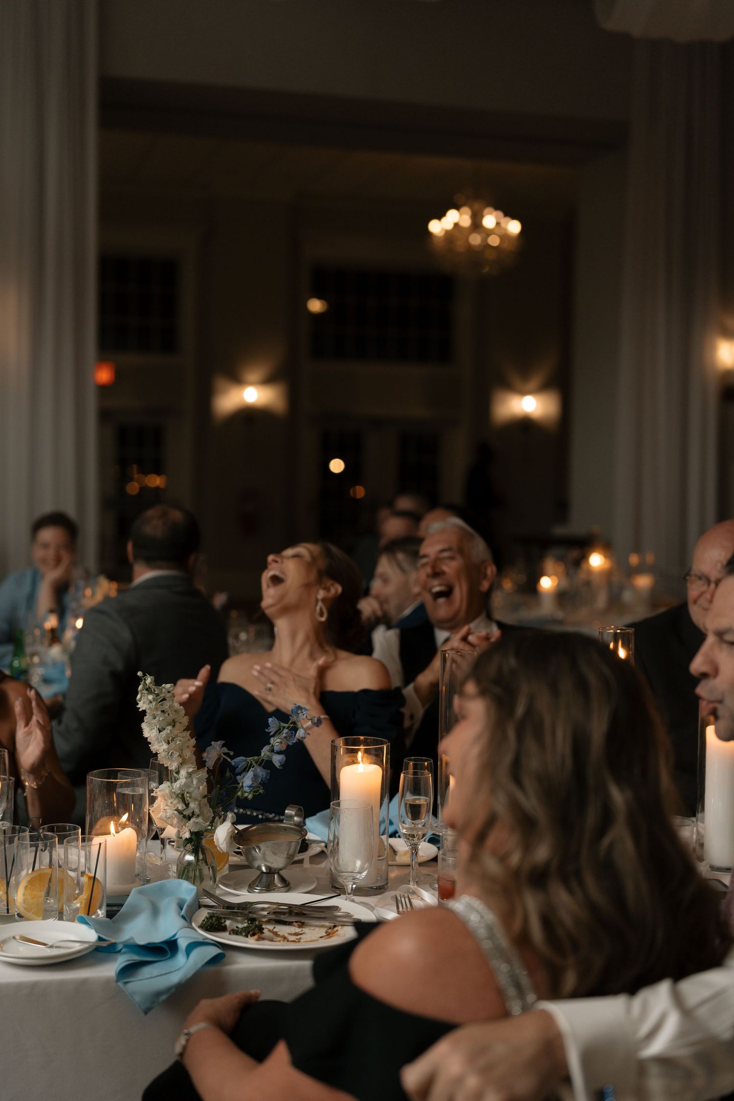Wedding guests laugh and celebrate at candlelit reception dinner table in elegant ballroom.