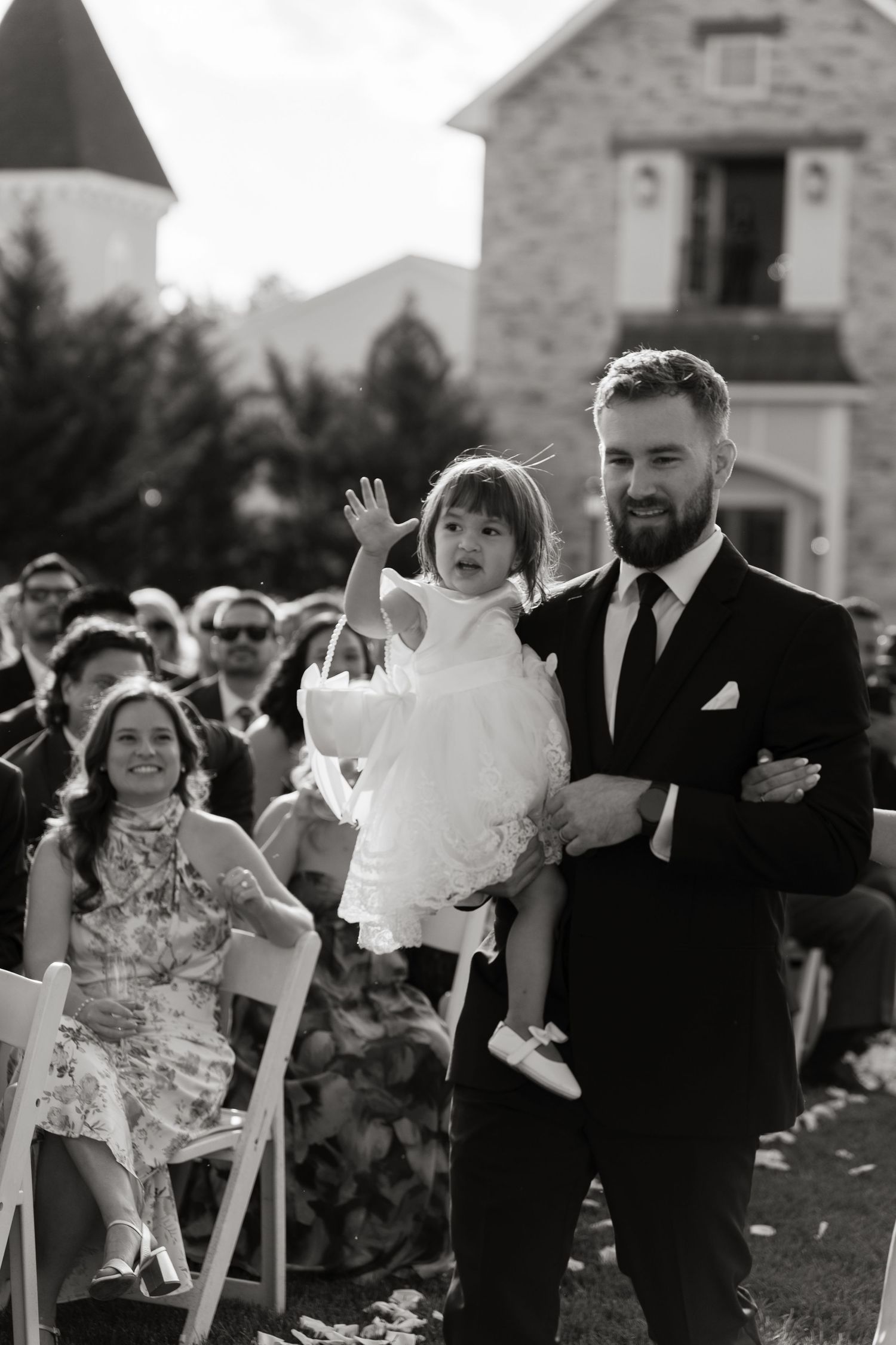 Black and white photo of bearded groomsman carrying flower girl down wedding ceremony aisle.