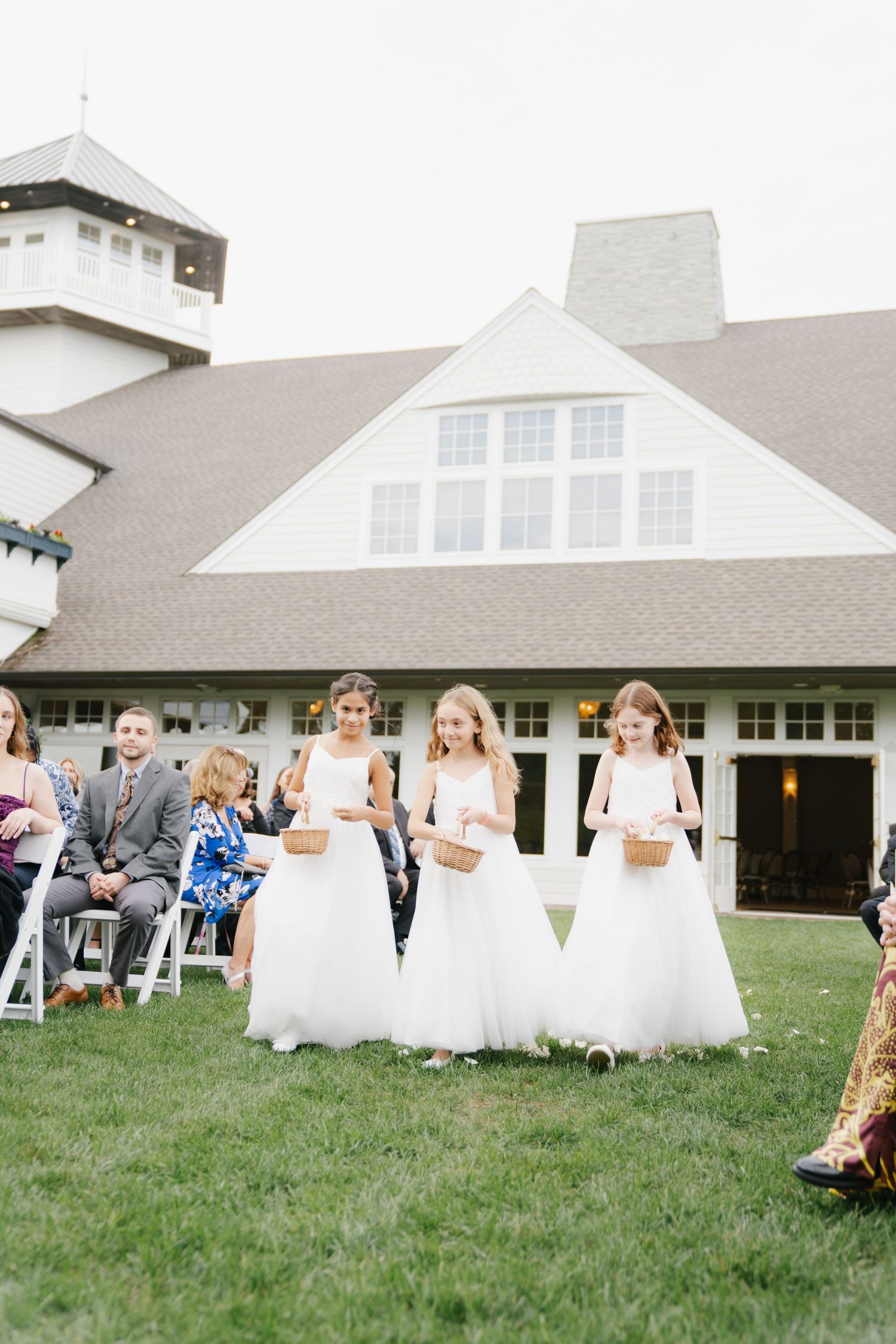 Young girls in white dresses run and play on a grassy lawn outside a white building during a wedding celebration.