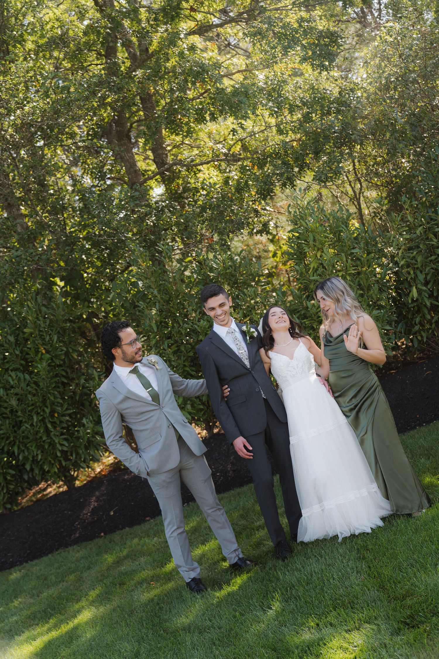 Wedding party laughs together on a sunny day in a garden setting with lush greenery in the background.