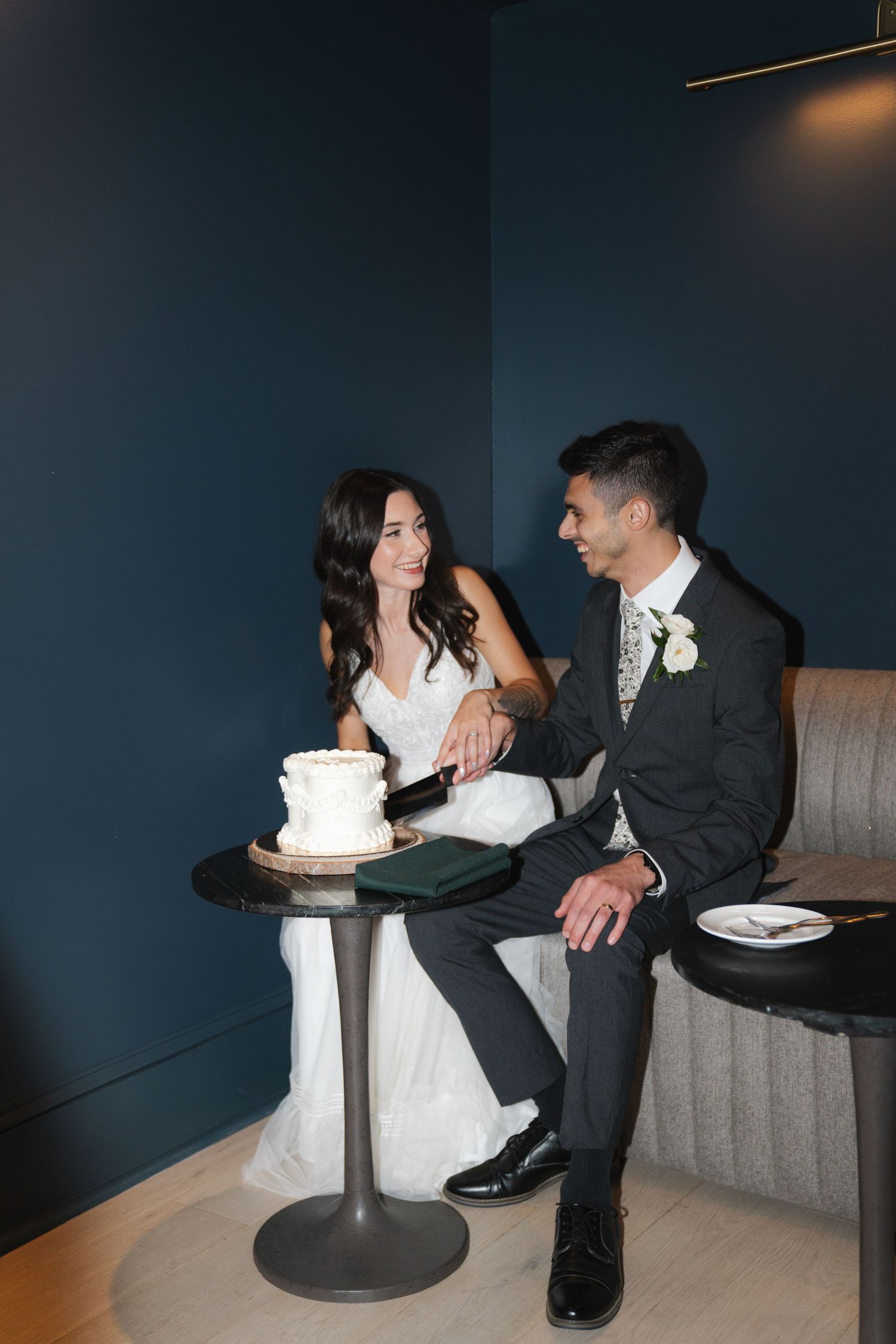 A couple sits together near a white wedding cake on a pedestal in a dimly lit room.