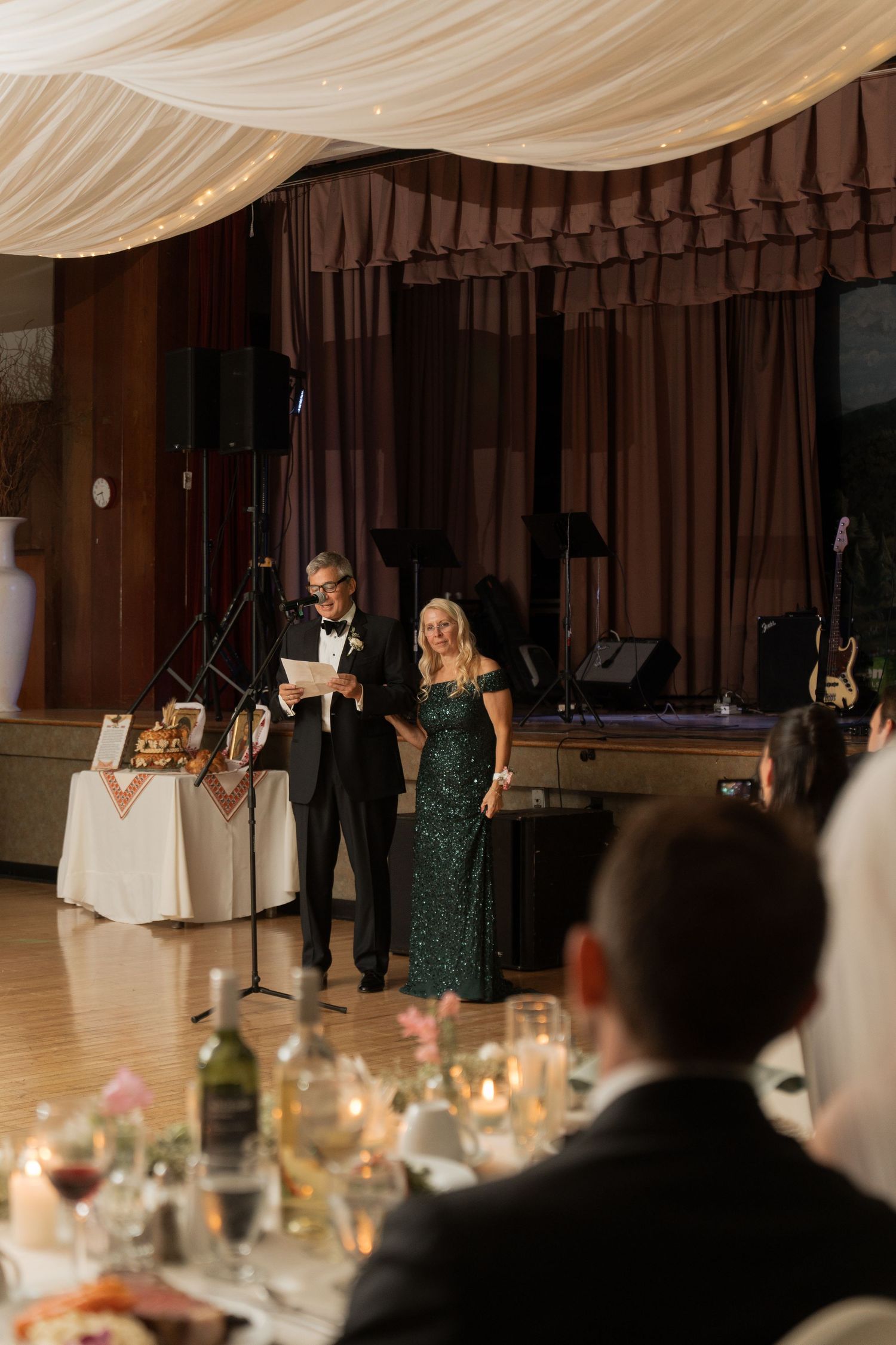 Couple in formal attire delivers heartfelt wedding toast speech on a decorated reception venue stage.