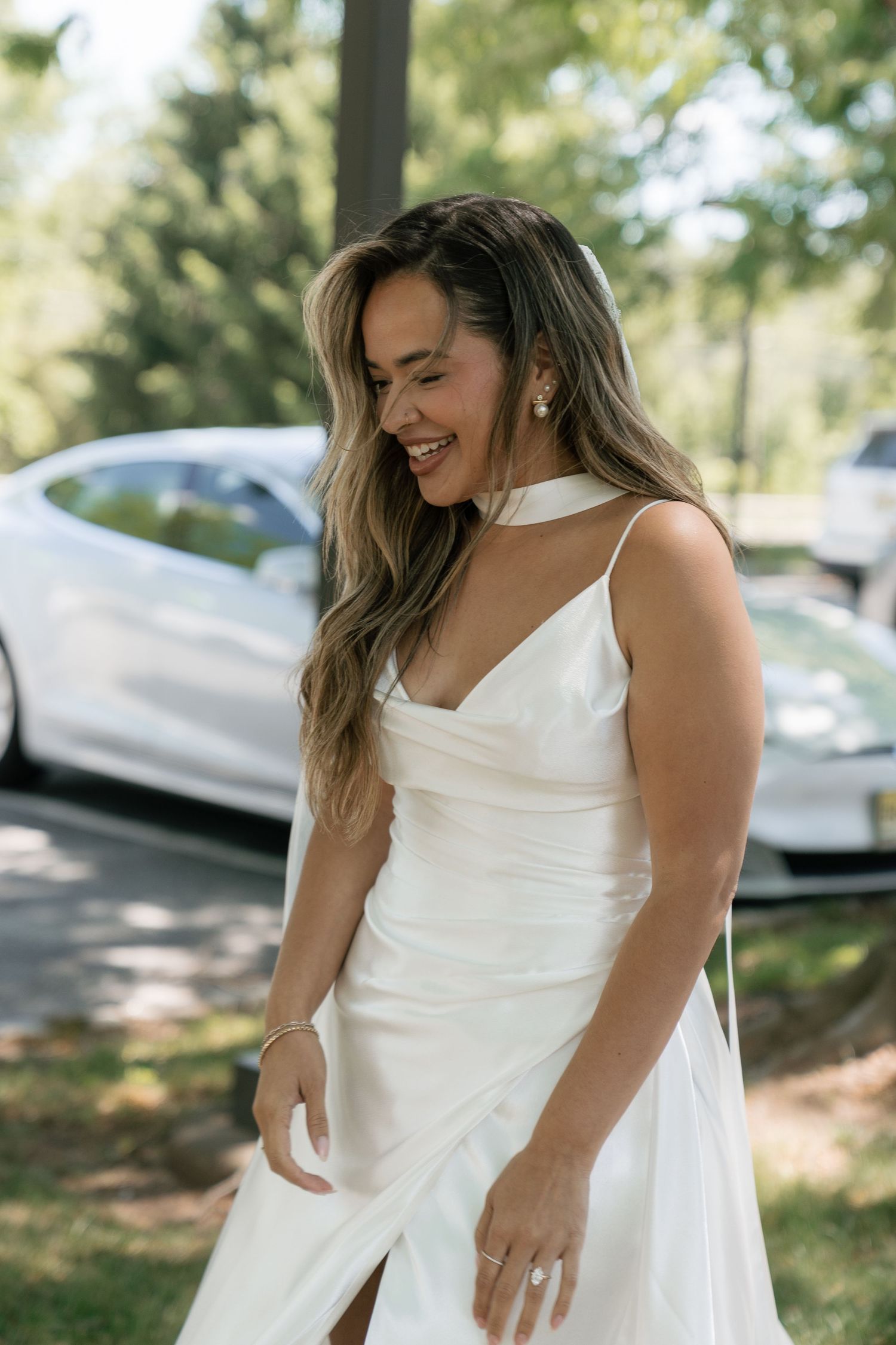 Smiling bride in elegant white satin slip wedding dress laughing outdoors near trees.