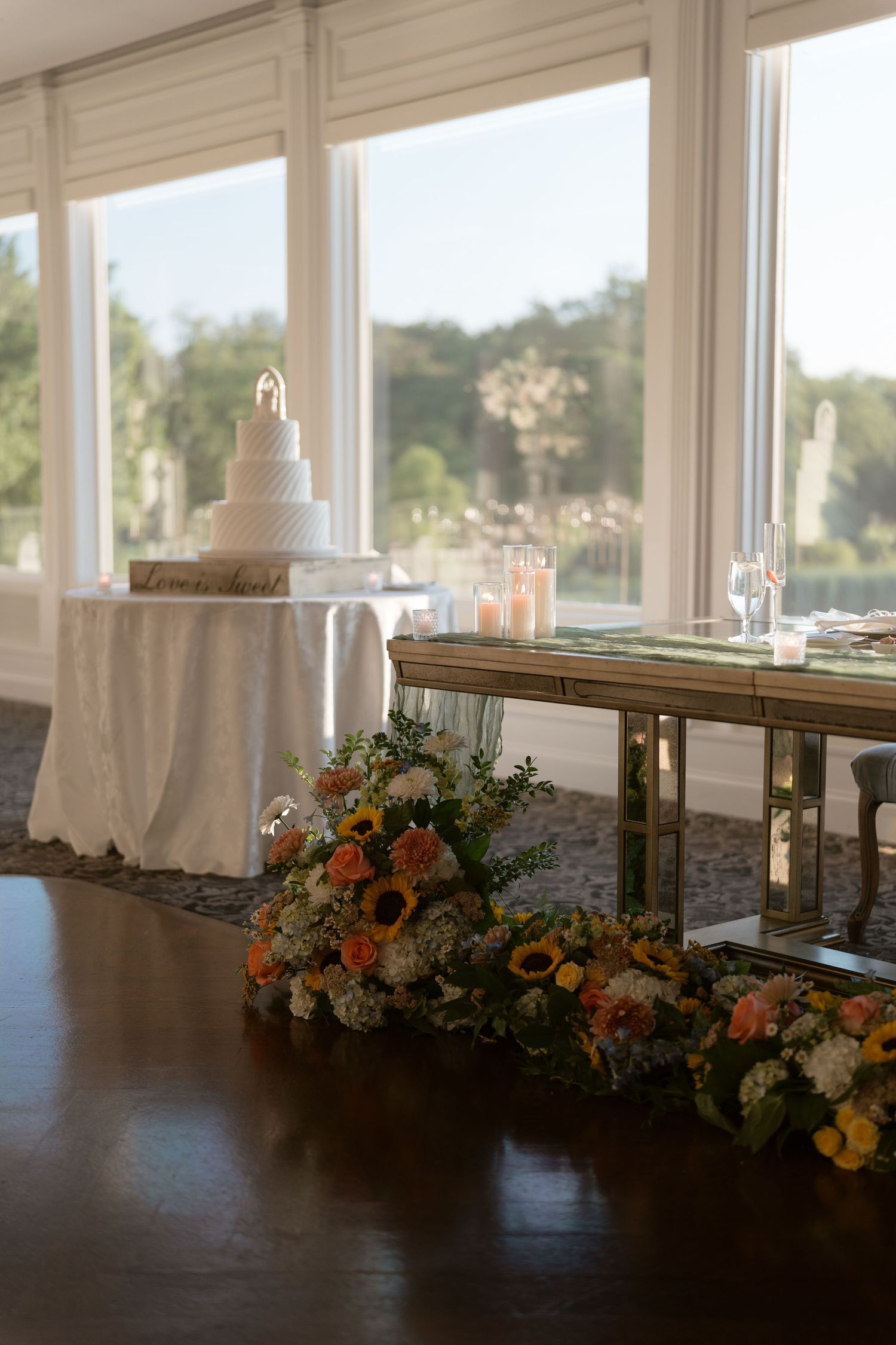 Elegant wedding reception setup with tiered white cake, floral arrangements of sunflowers, and candles by large windows.
