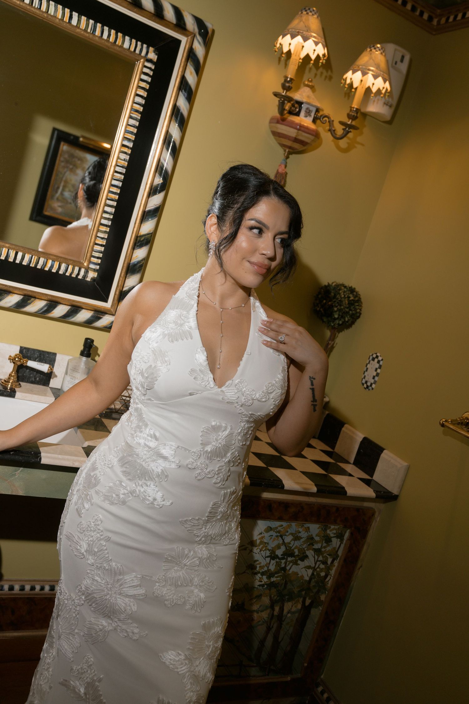 Bride in a white halter lace wedding gown poses elegantly in a decorated bathroom with ornate mirror.