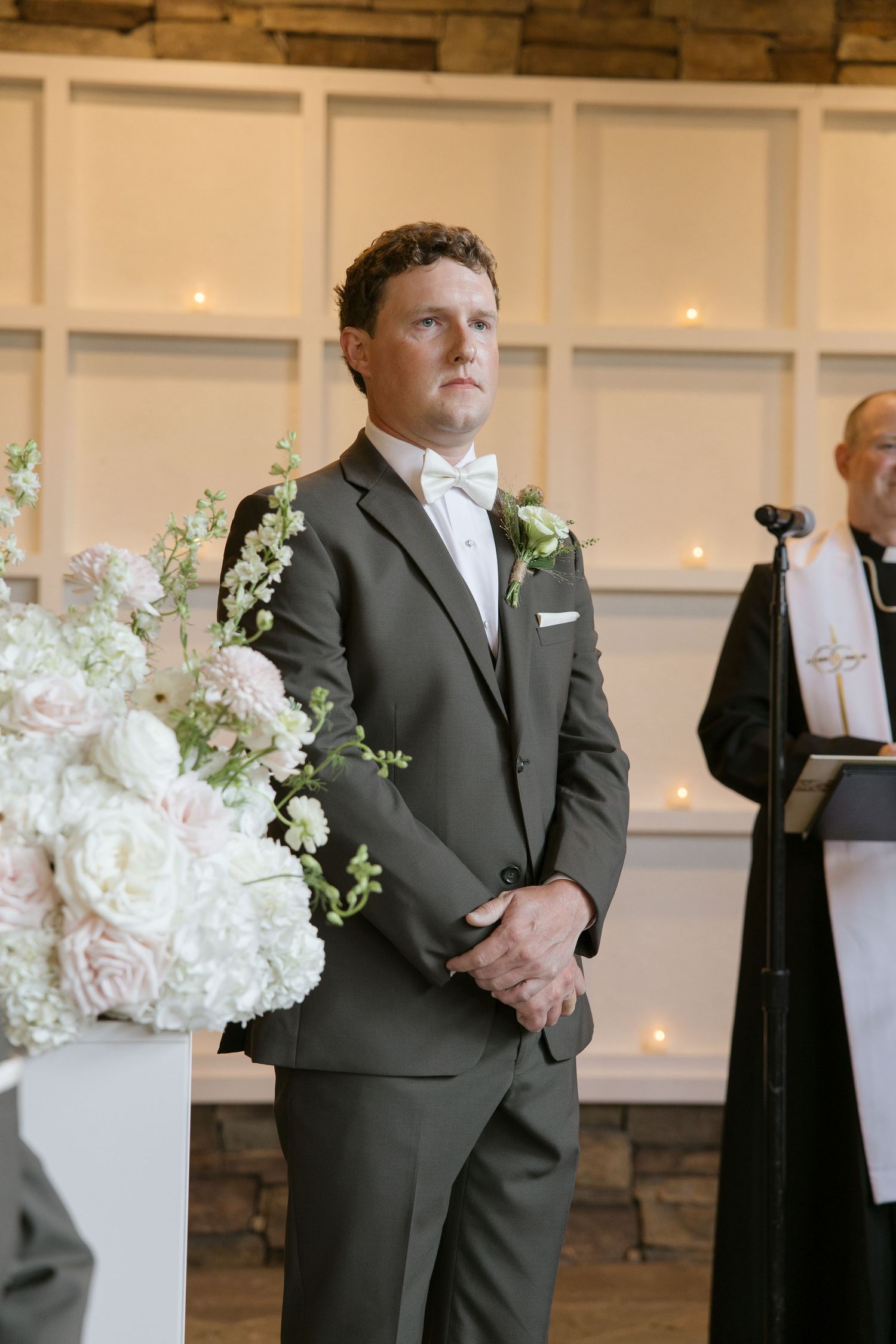 Groom in gray suit and white bow tie stands at altar with floral arrangements during wedding ceremony.