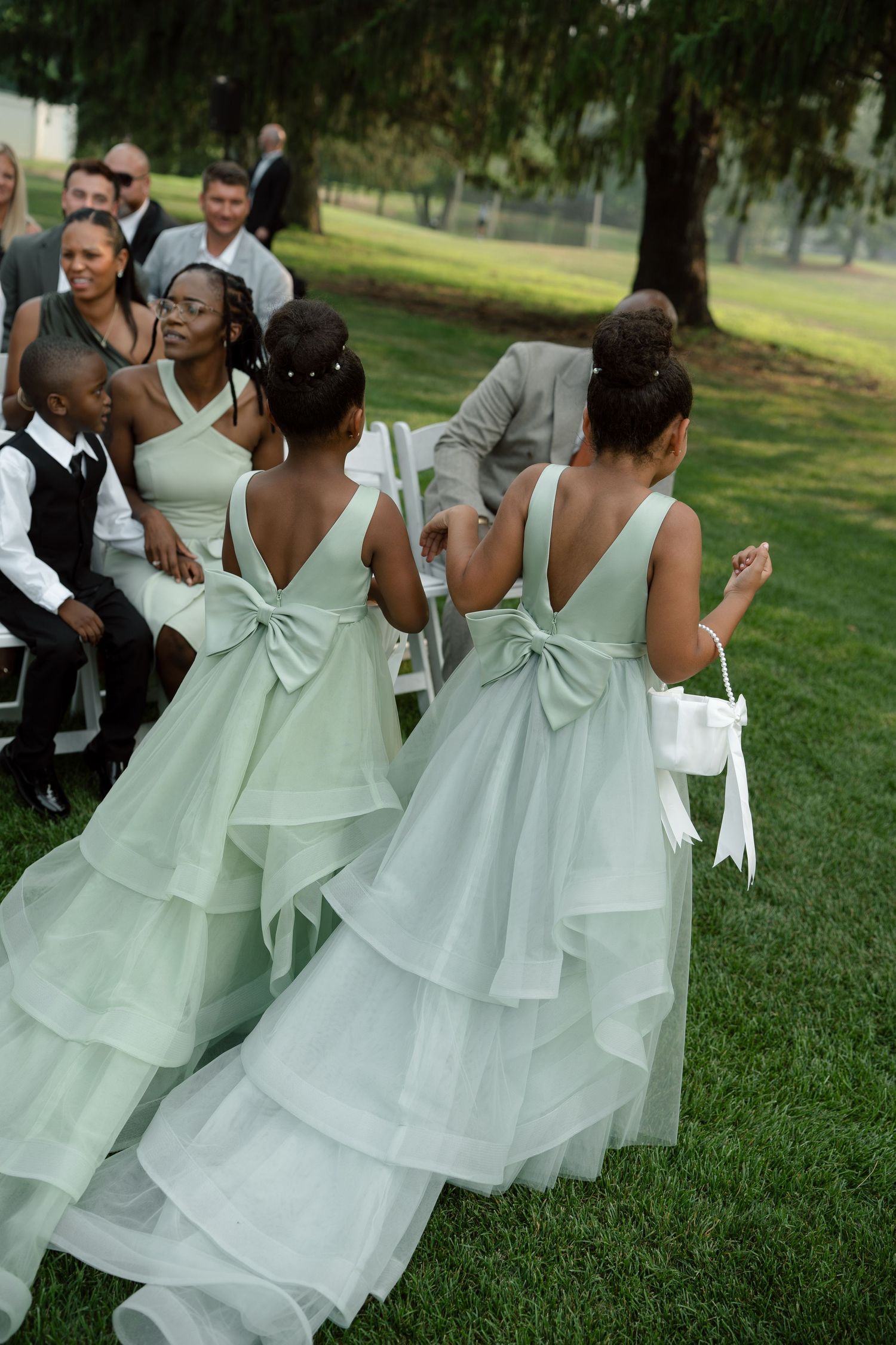 Two flower girls in sage green tiered gowns with bows walk down outdoor wedding aisle.
