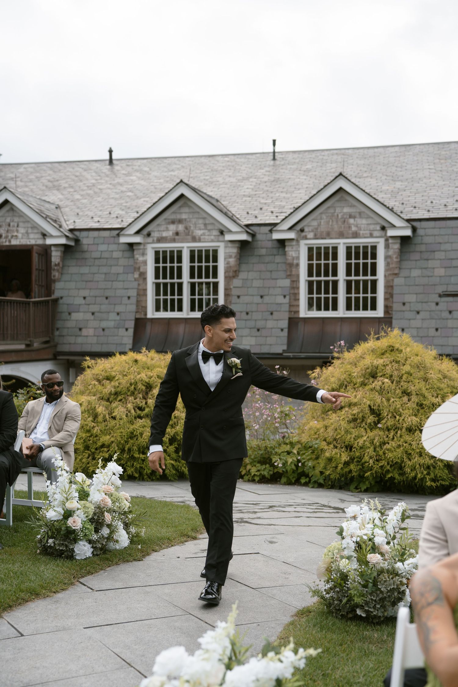 Groom in black tuxedo walks down outdoor wedding aisle lined with white floral arrangements at rustic venue.