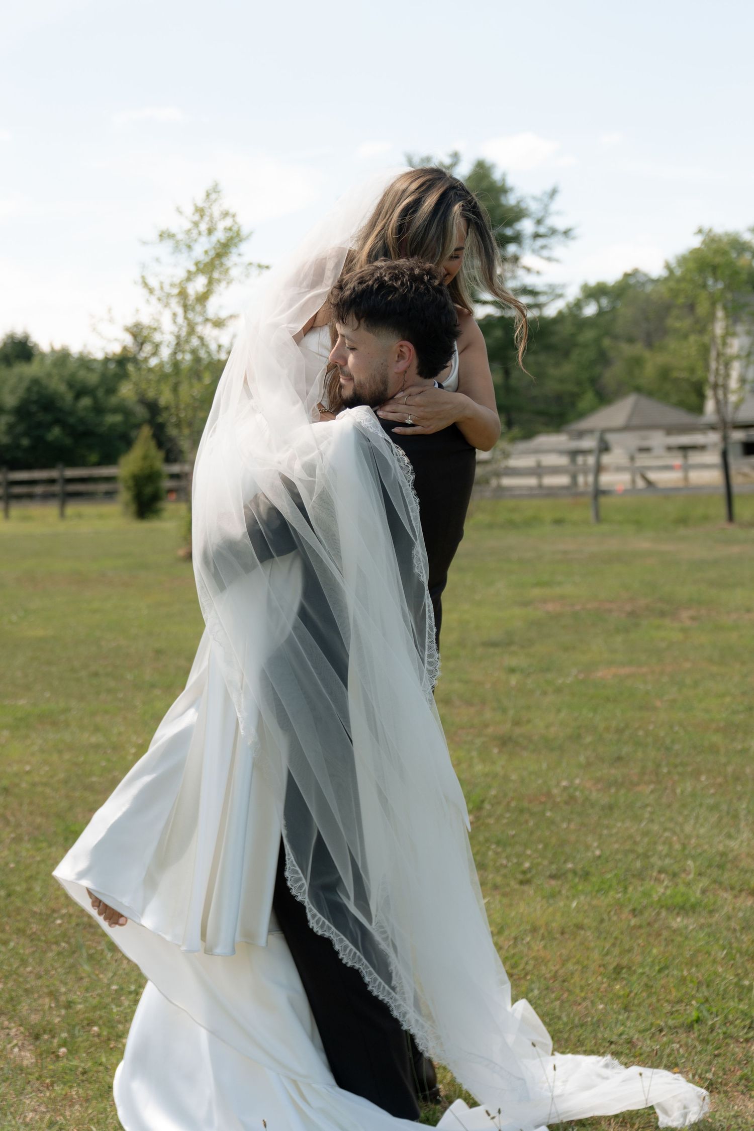 Groom lifting bride in white wedding gown and veil in green outdoor field on wedding day.