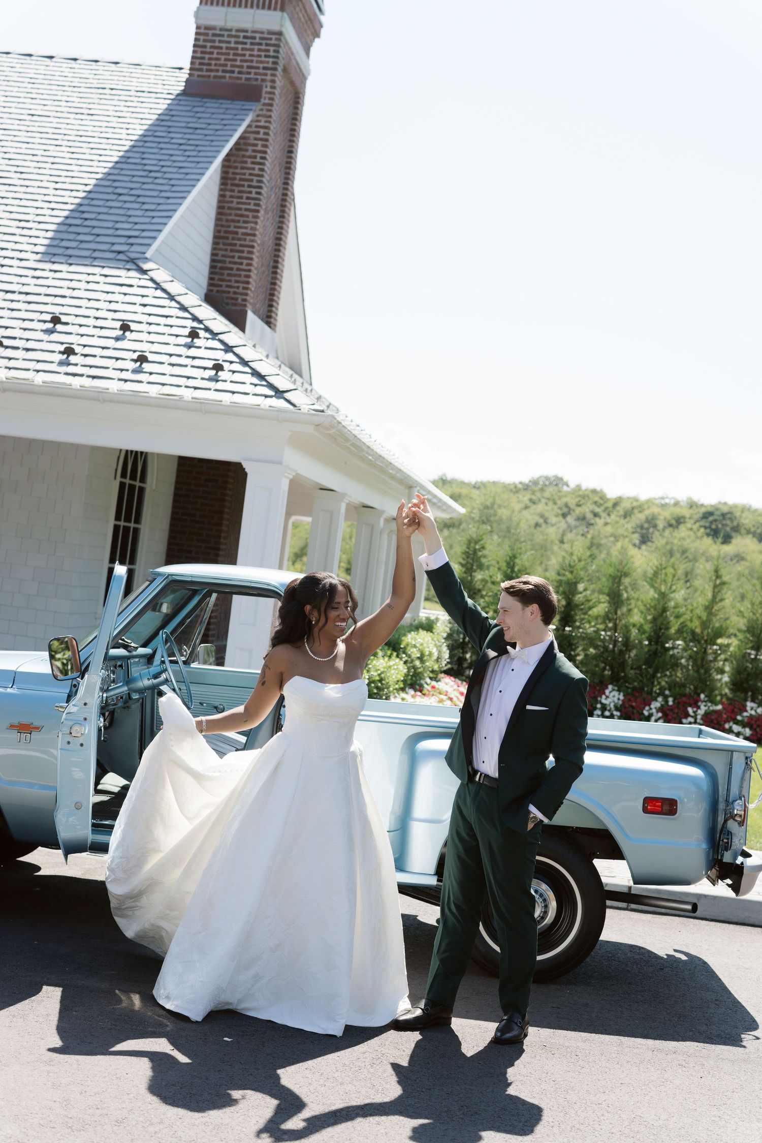 Bride and groom dancing joyfully in front of a vintage light blue truck outside a charming wedding venue.