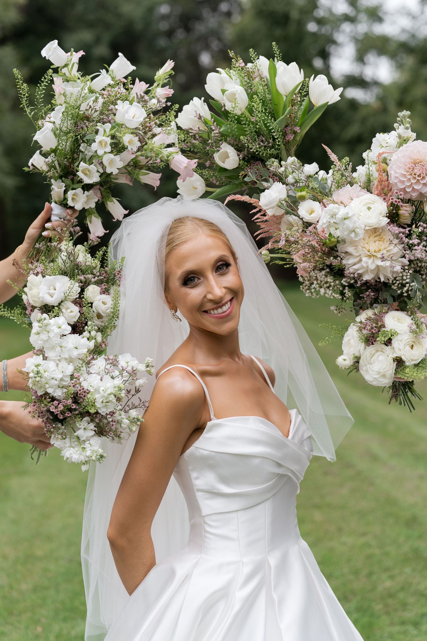 Smiling bride in white satin gown surrounded by elegant white and pink floral bouquets outdoors.