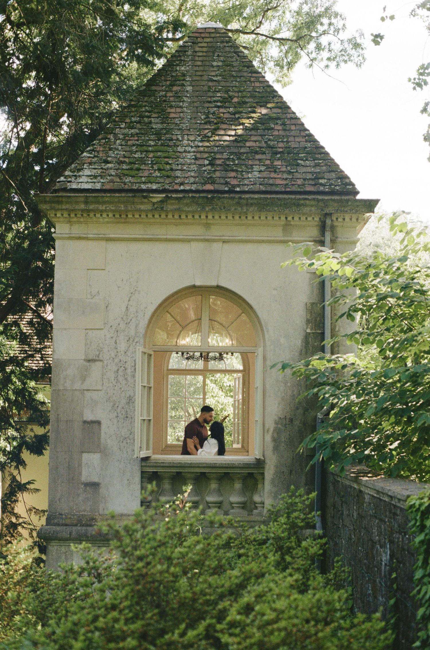 Couple sharing a romantic moment in an arched window of a classic stone garden pavilion surrounded by lush greenery.