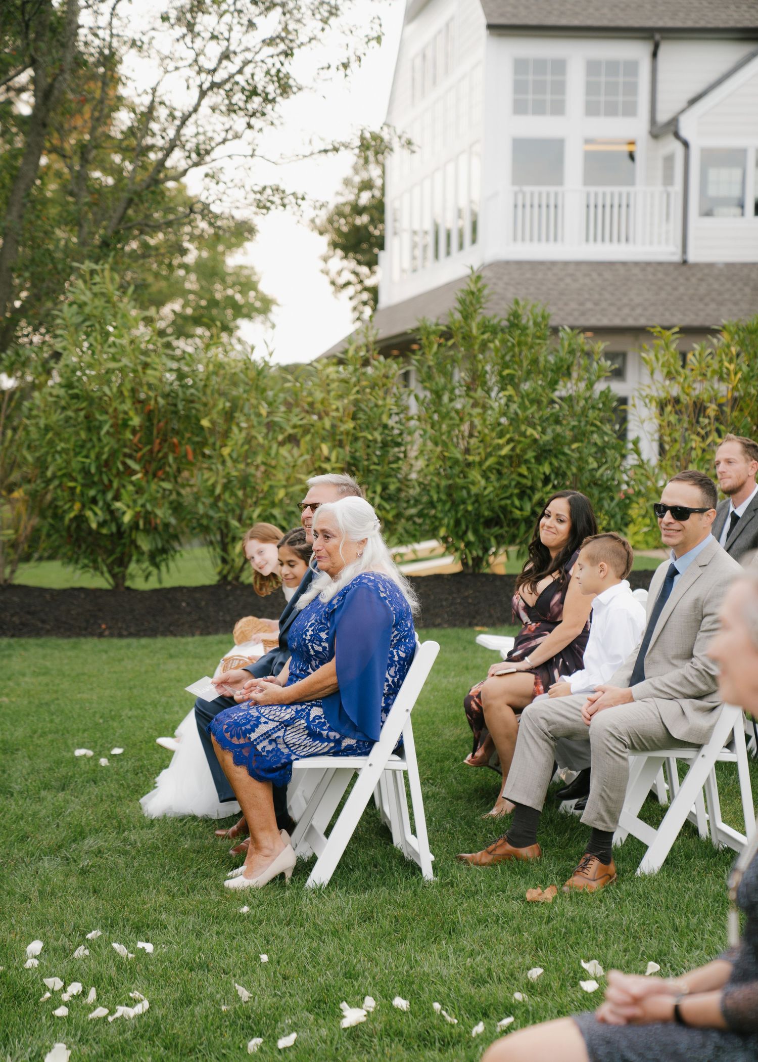Wedding guests relax in white chairs on a grassy lawn during an outdoor celebration.