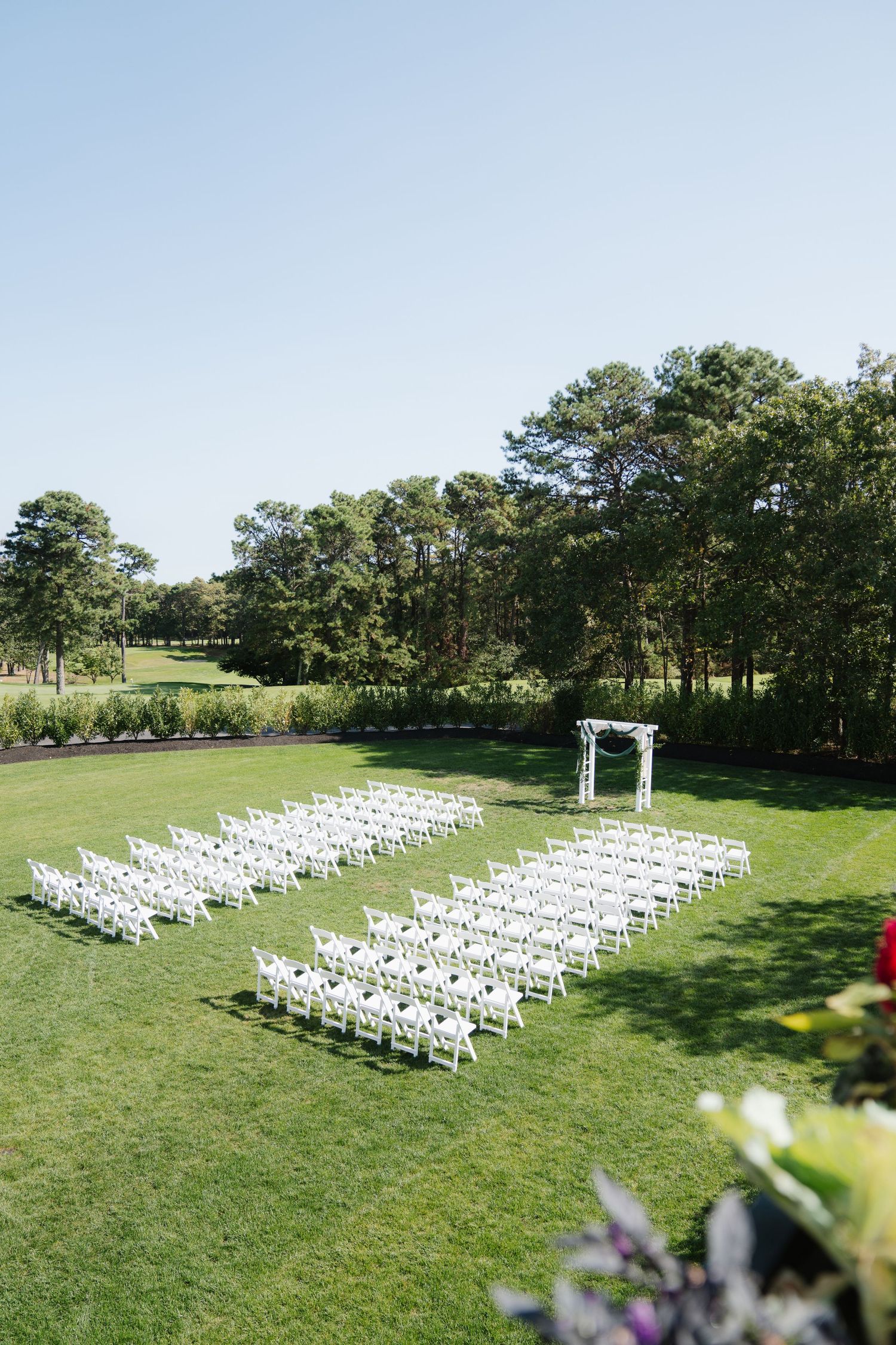 White chairs arranged in rows on a manicured lawn for an outdoor wedding ceremony with an arch under tall pine trees.