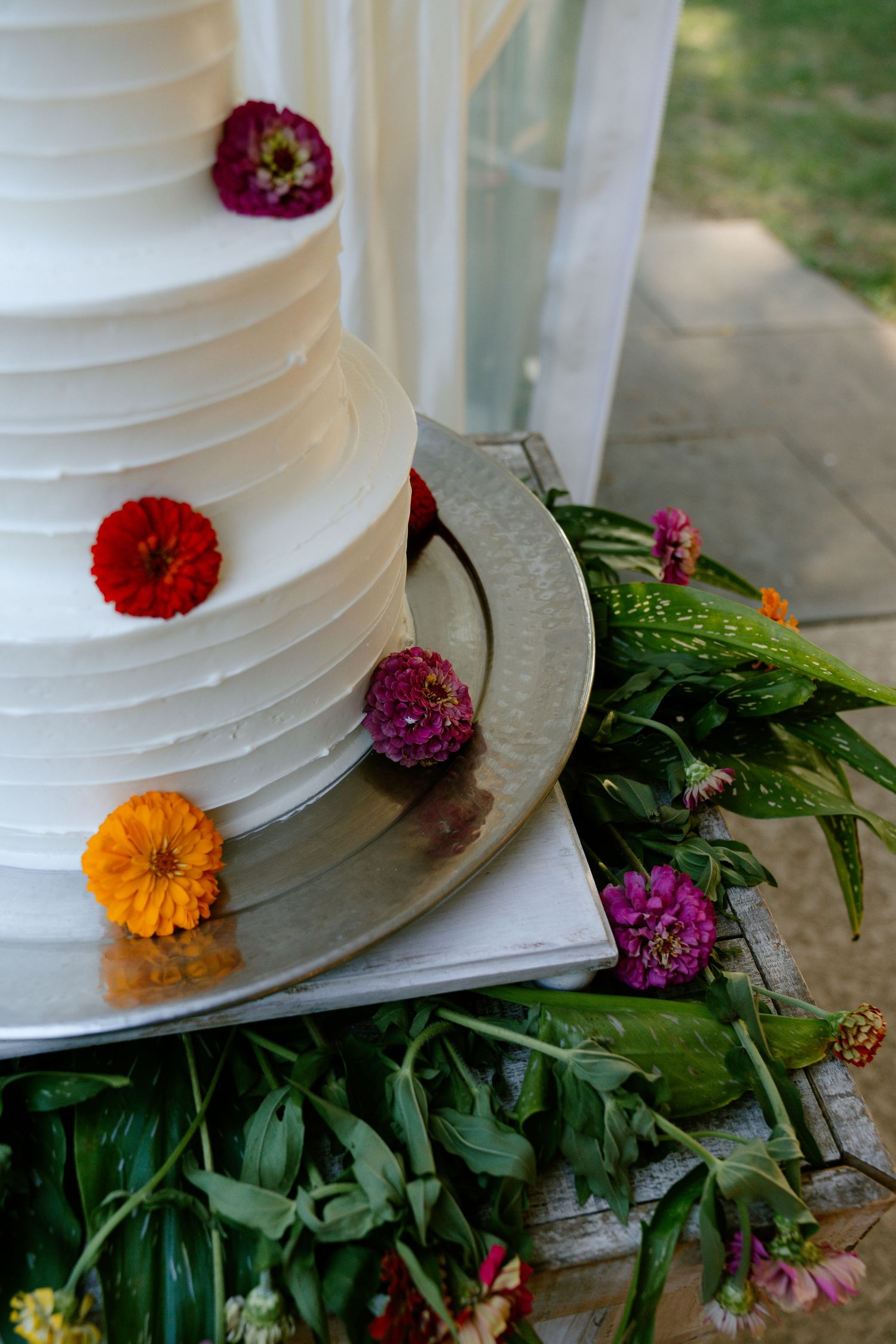 White wedding cake decorated with colorful fresh flowers and greenery on rustic wooden surface.