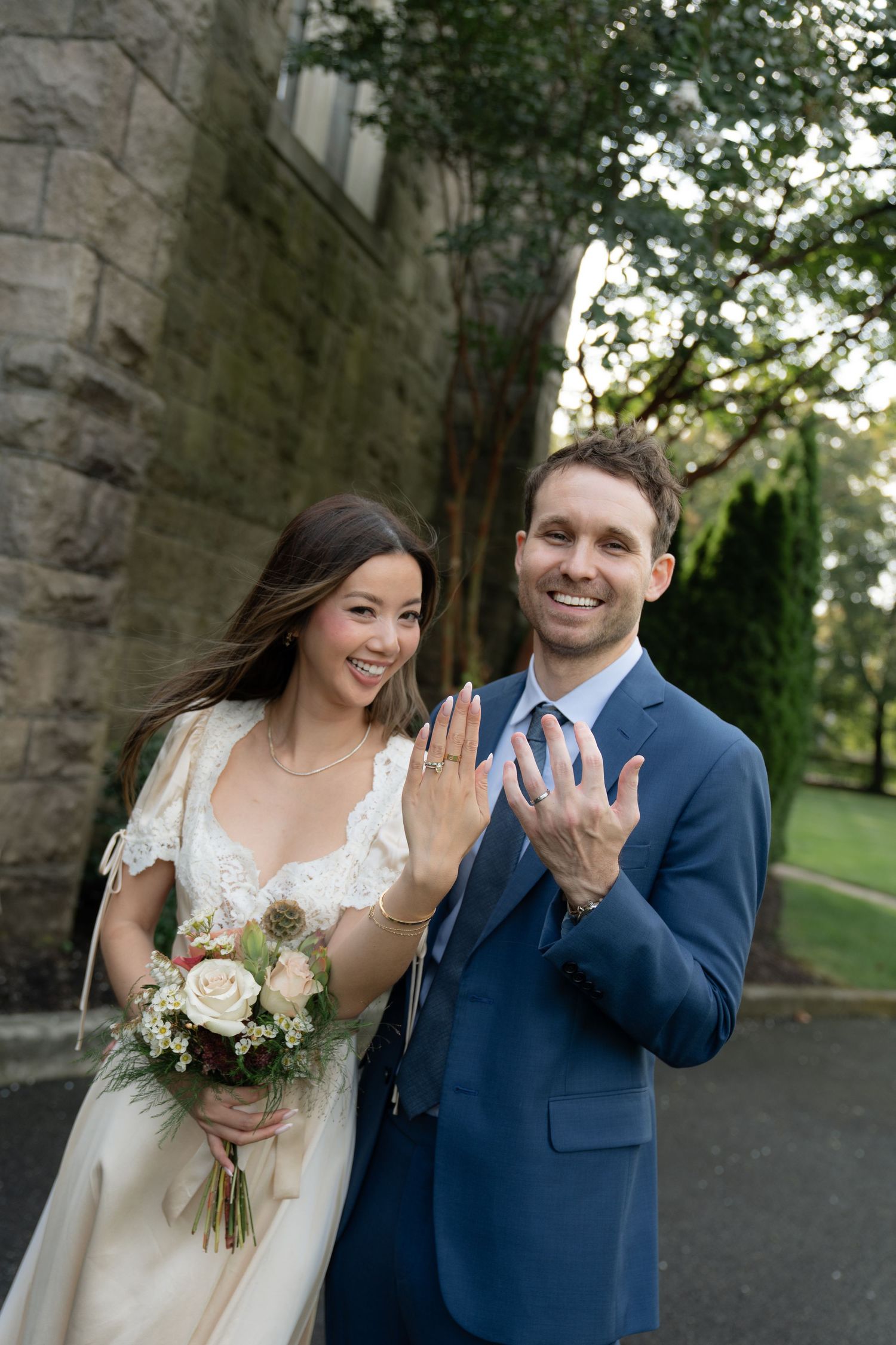 A happy newlywed couple in formal wedding attire poses together showing off their rings against a stone wall background.