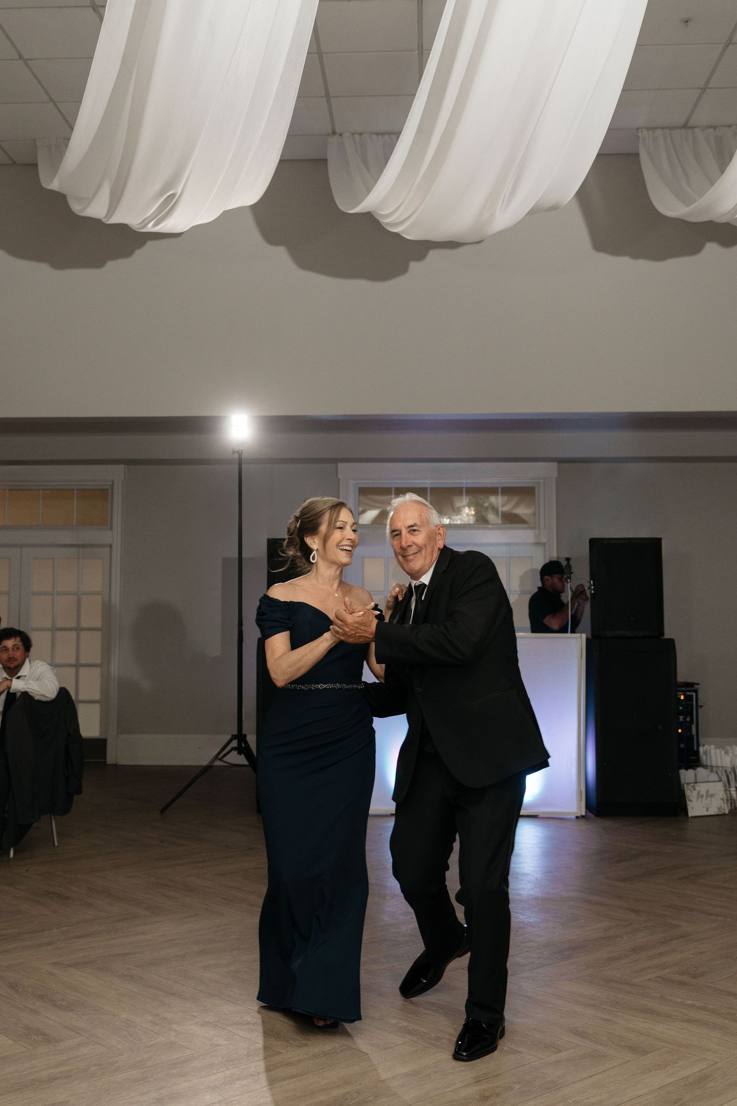 Elegantly dressed couple sharing a joyful dance at a wedding reception with draped ceiling.