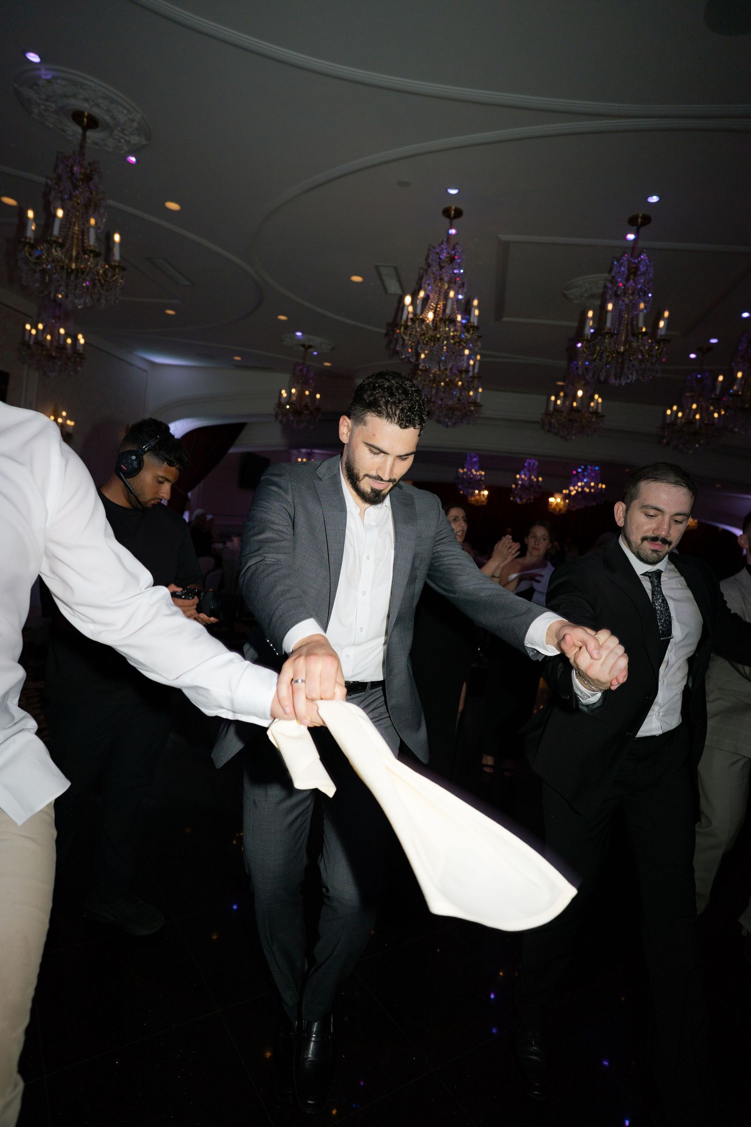 Wedding guests dancing and pulling napkins on a dark dance floor under crystal chandeliers.