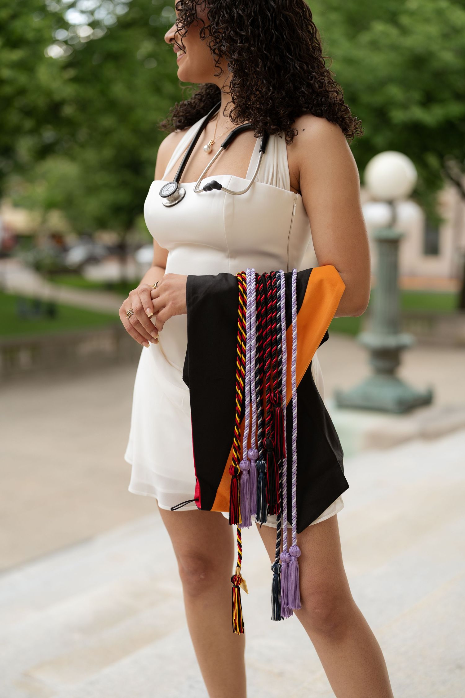 Nursing graduate in white dress holding colorful graduation stoles and cords with stethoscope outdoors.