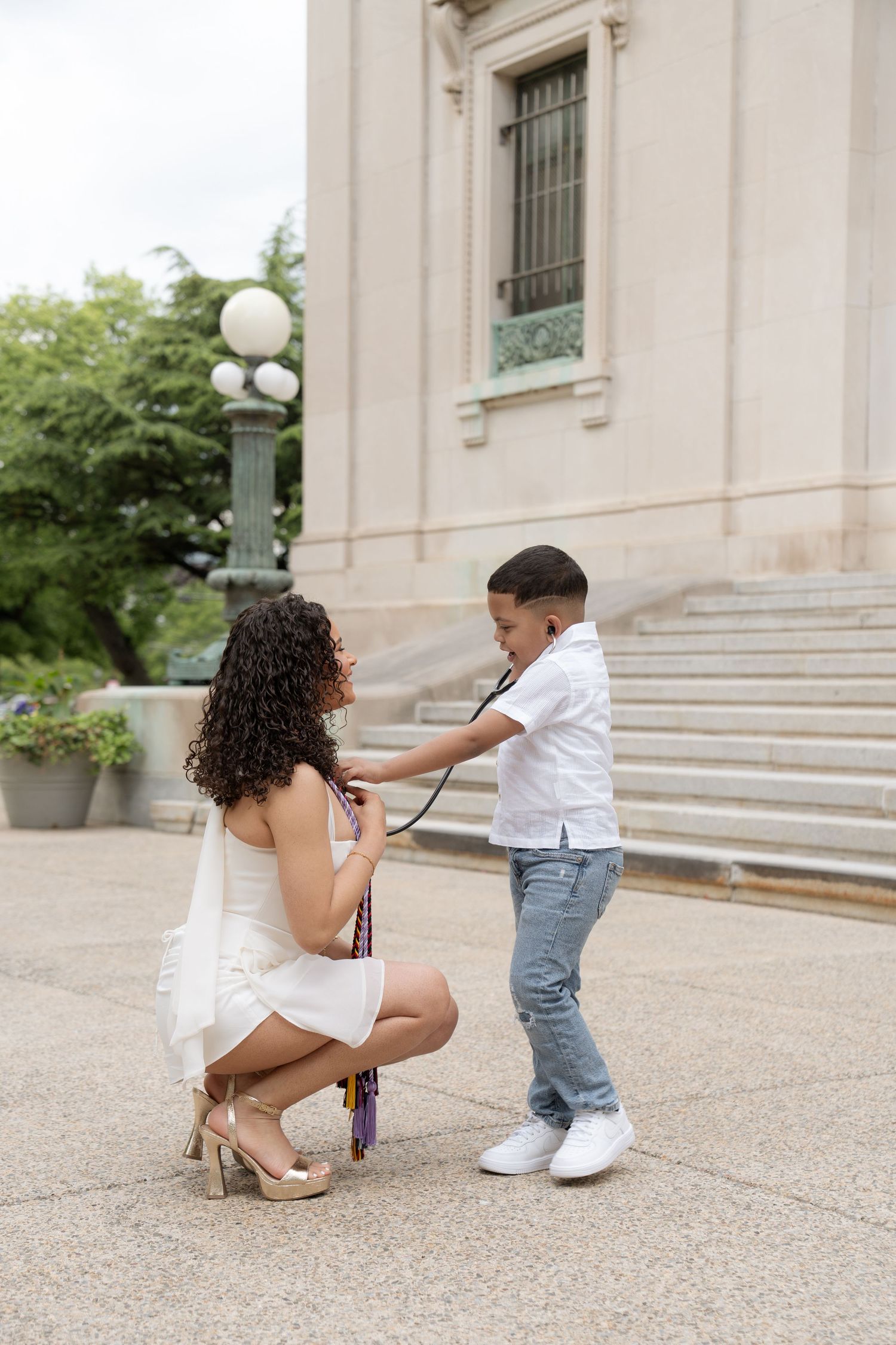 A young boy uses a stethoscope on a woman in a white dress crouching outdoors near a stone building.