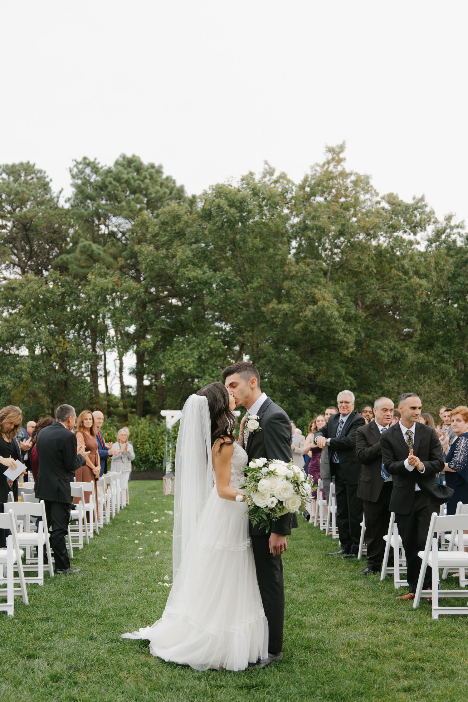 Wedding ceremony on a grassy slope with white chairs and guests gathered under green trees at sunset.