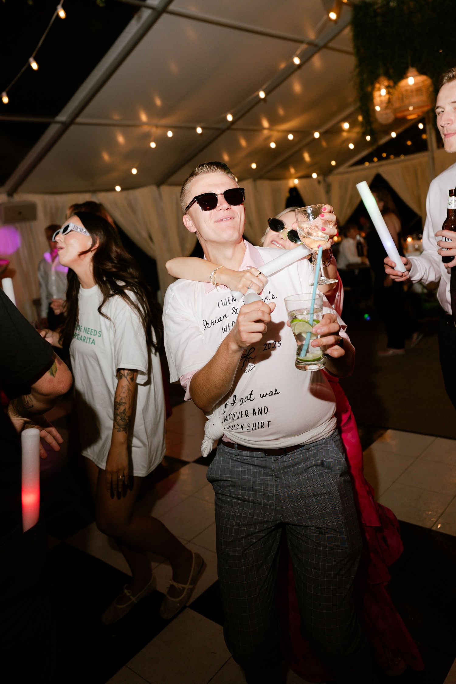People dancing and holding drinks at a party under string lights in a dimly lit indoor venue.