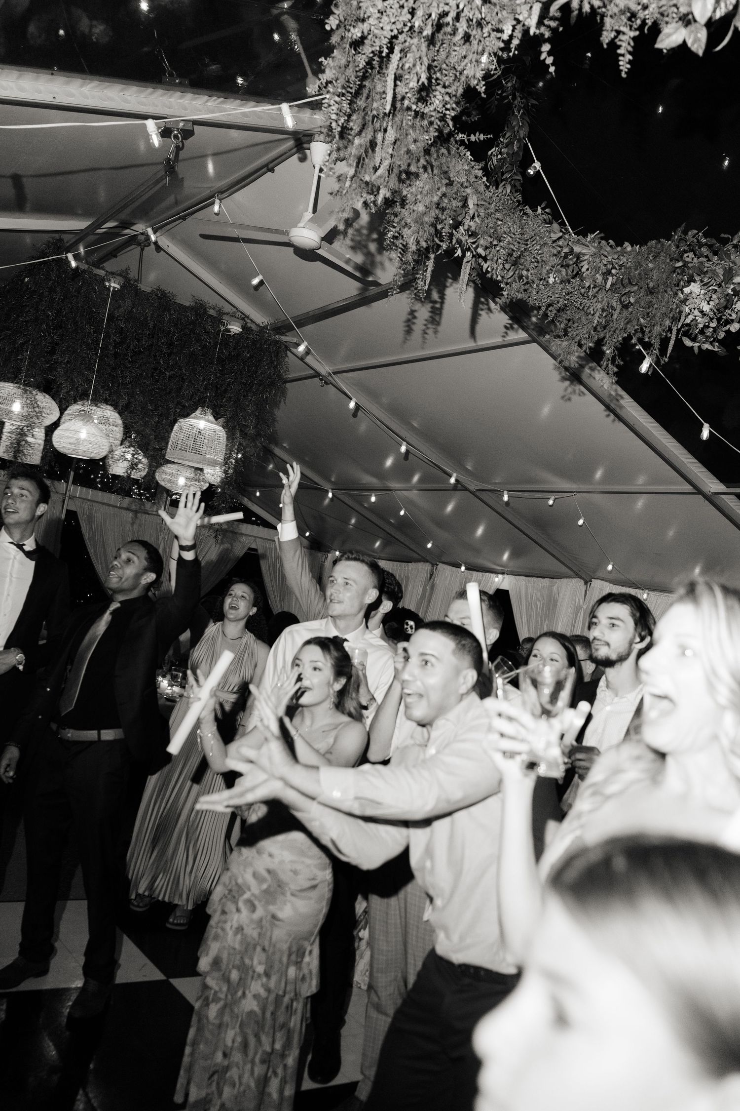 Black and white photo of wedding guests dancing and celebrating under string lights at an evening reception.