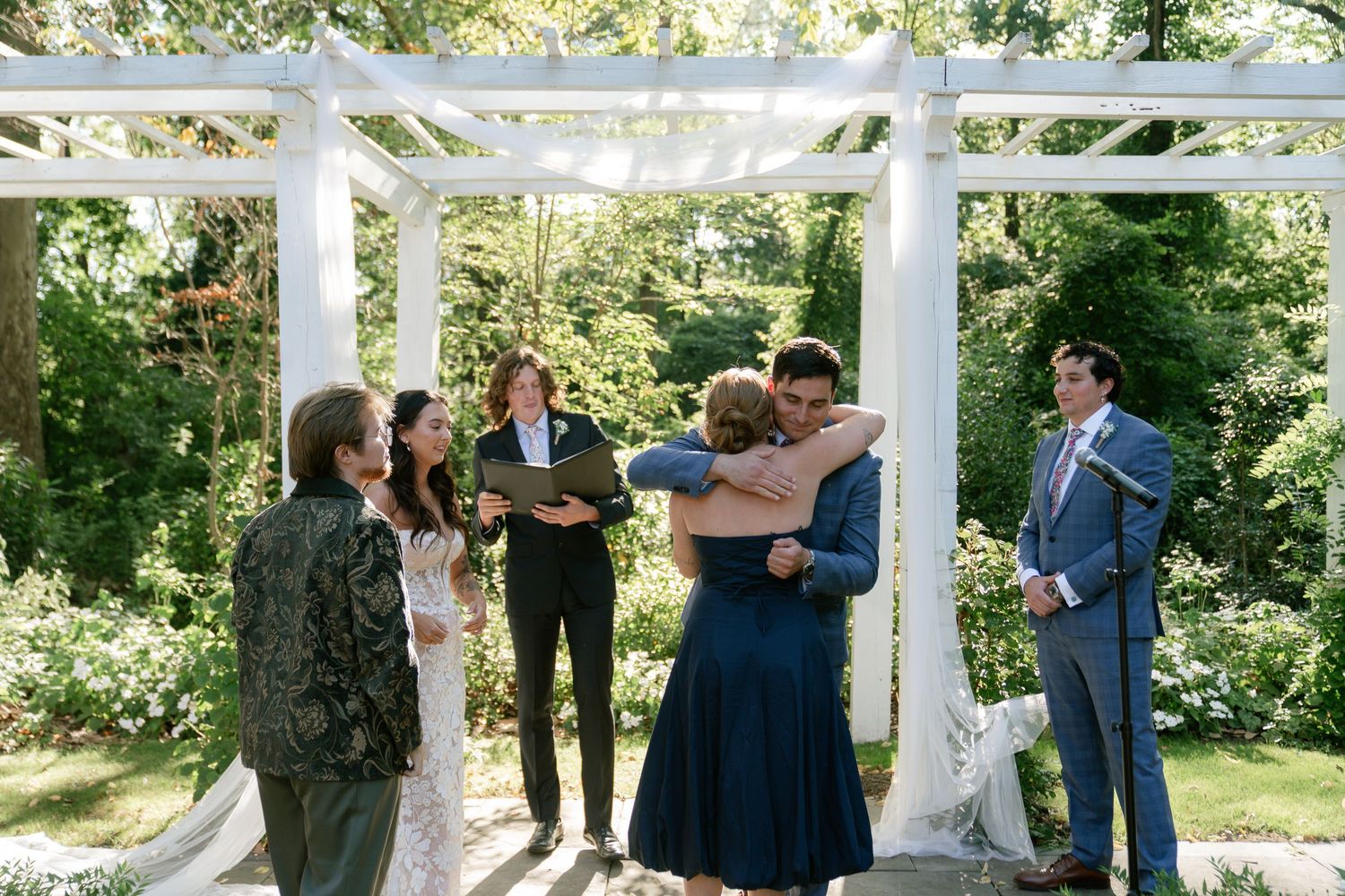 Outdoor wedding ceremony taking place under a white gazebo surrounded by lush greenery and trees.